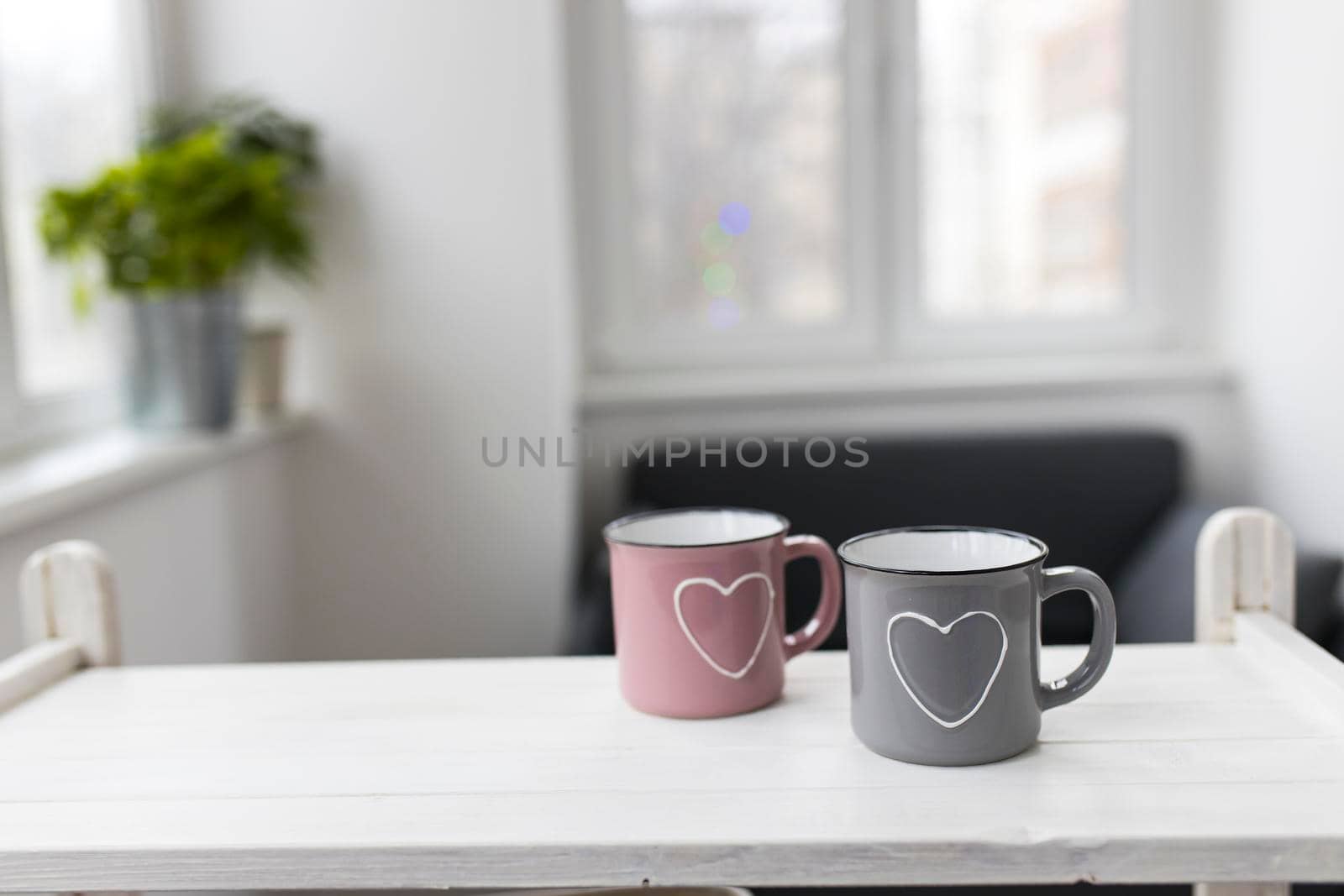 Gray and pink with hearts enamel cups sit on the table in front of the window. Valentine's Day.