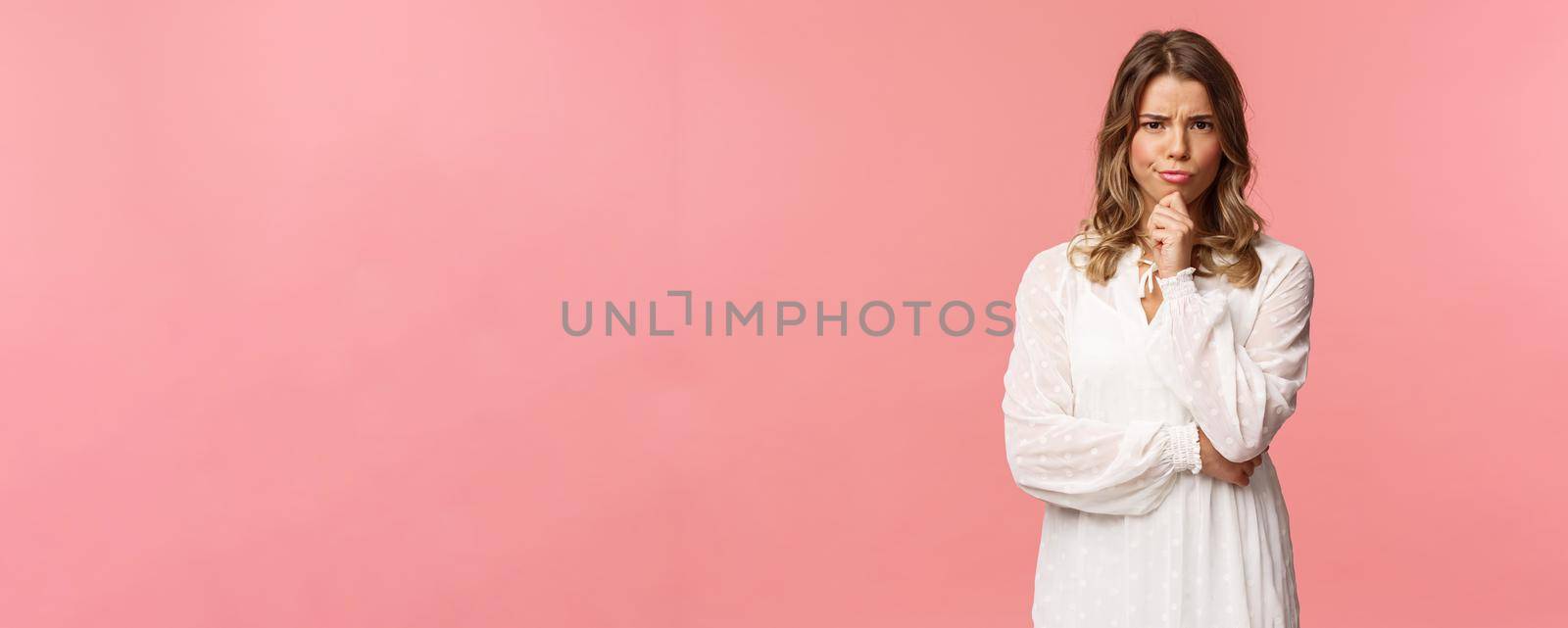Beauty, fashion and women concept. Portrait of skeptical and judgemental serious-looking blond female in white spring dress, look disbelief, grimacing and frowning, pink background.