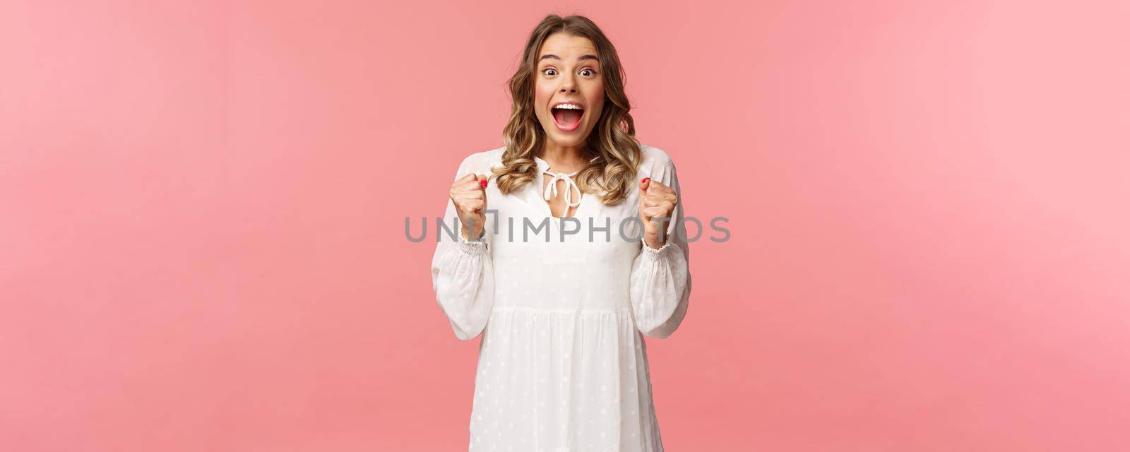 Portrait of surprised lucky hopeful, blond girl winning, hear great news and chanting, gasping open mouth smiling joyful, jumping from happiness and joy, standing pink background.