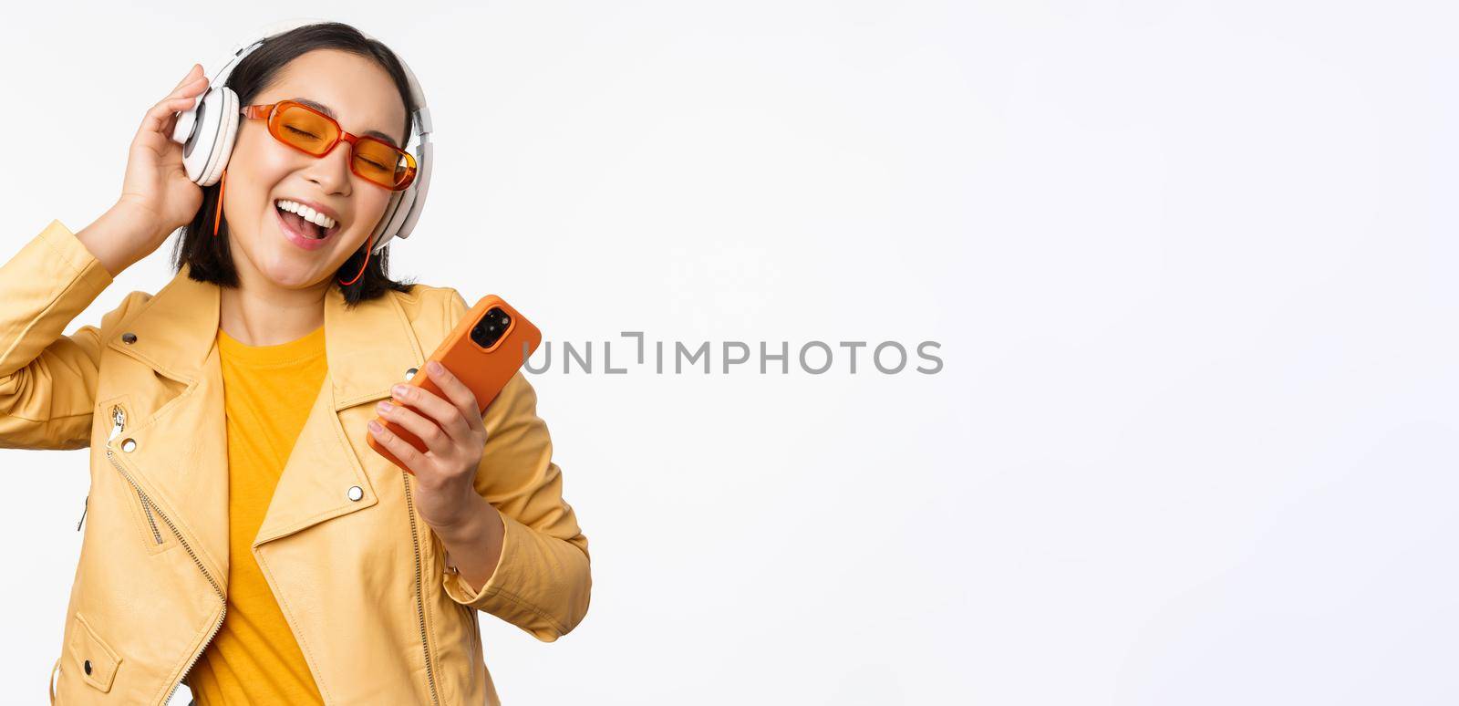 Technology concept. Stylish asian girl in headphones, holding smartphone, dancing and singing, listening music, standing over white background.