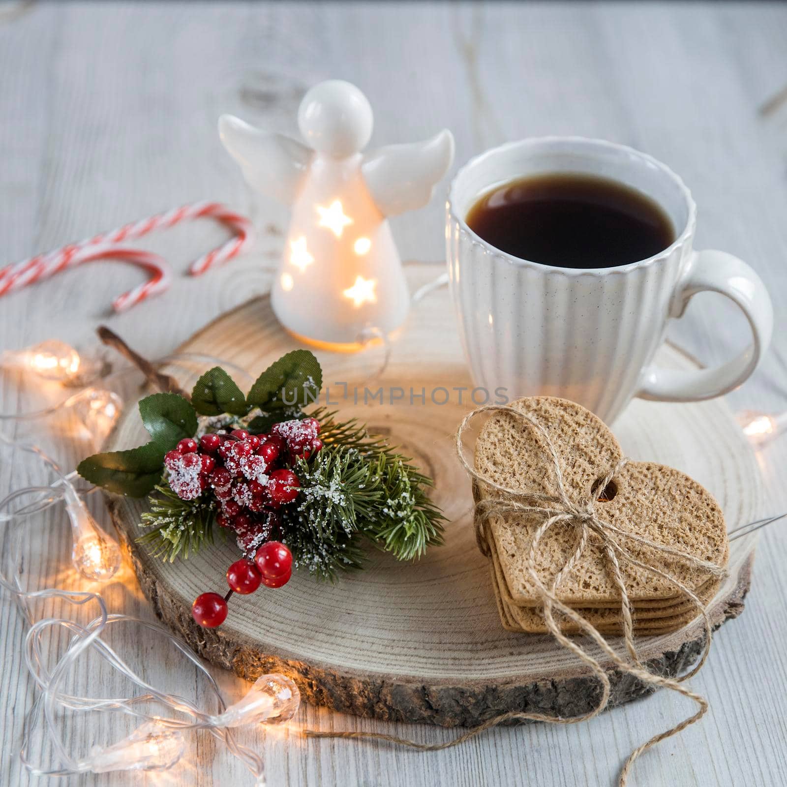 A cup of coffee on a wooden stand, a gingerbread cookie tied with a rope, a garland on the table. Artificial spruce branch with red berries. Breakfast at Christmas. Scandinavian style. Copy space by elenarostunova