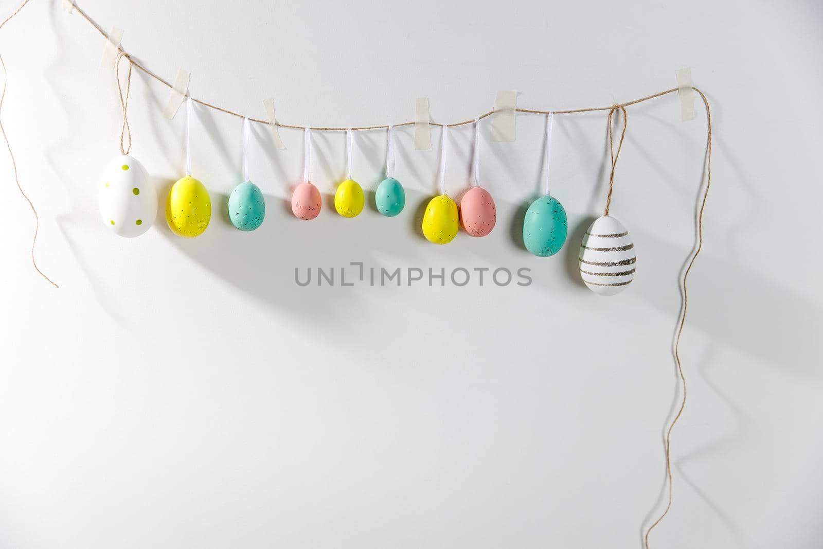 A garland of multi-colored plastic eggs hanging on a white wall. Home decoration for Easter by elenarostunova