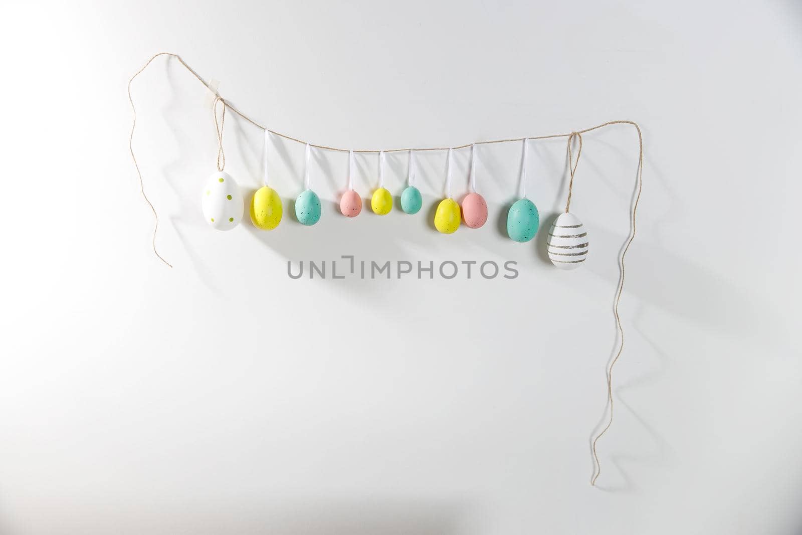 A garland of multi-colored plastic eggs hanging on a white wall. Home decoration for Easter