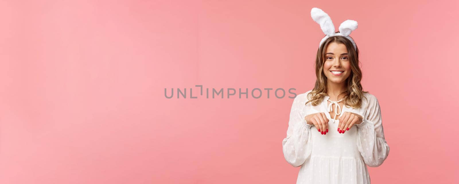 Holidays, spring and party concept. Portrait of cute and tender, lovely blond girl imitating bunny, holding hands like paws and wearing rabbit ears, smiling camera, pink background.