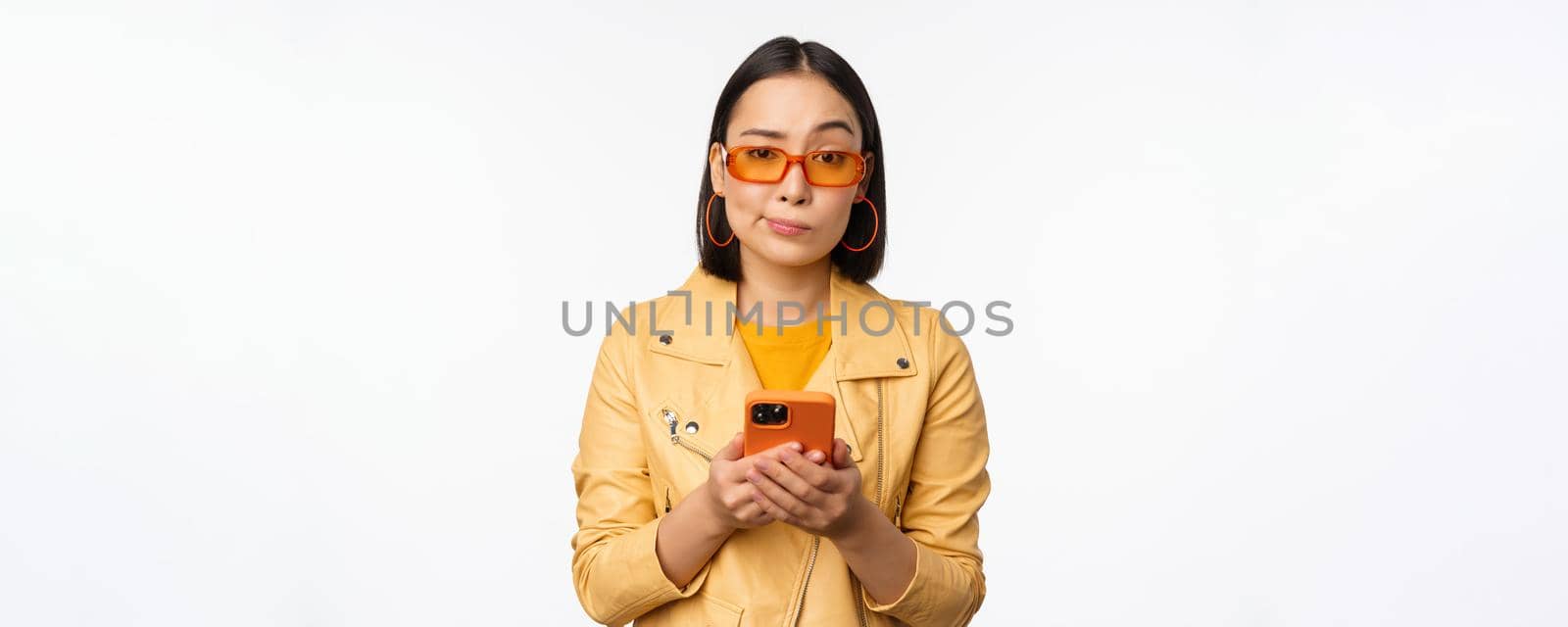 Portrait of confused asian girl in sunglasses, holding smartphone, using mobile phone and looking puzzled, standing clueless over white background.