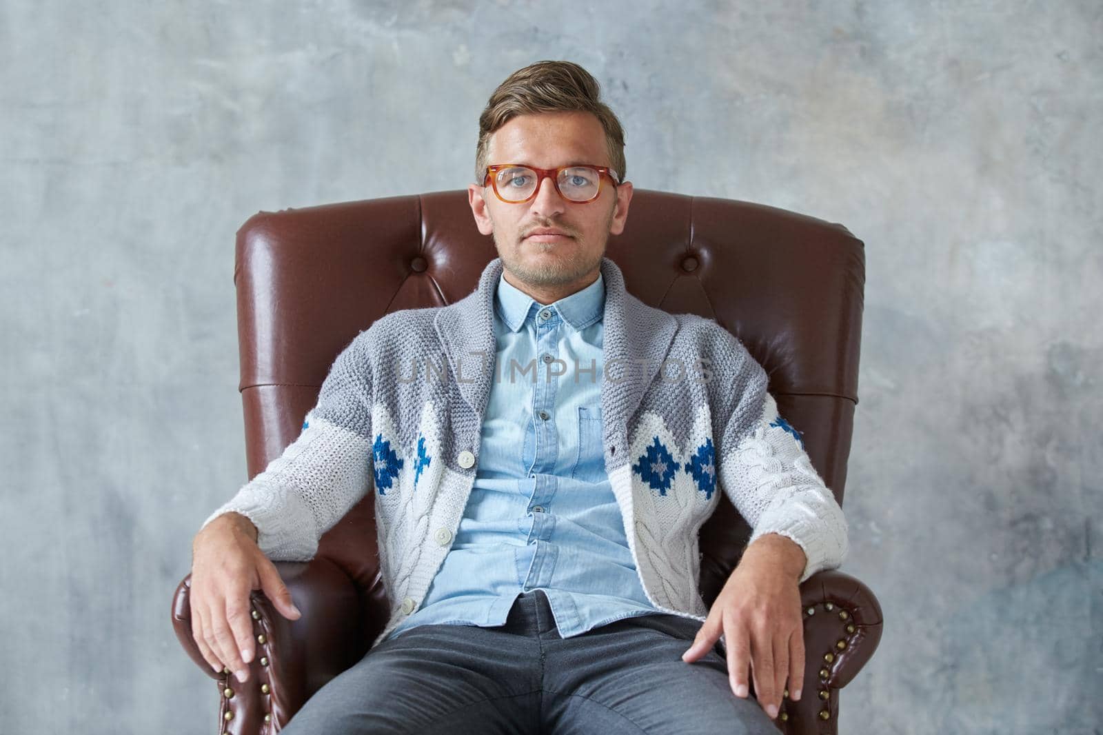 Portrait of a stylish intelligent man with glasses stares into the camera, good view, small unshaven, charismatic, blue shirt, gray sweater, sitting on a brown leather chair. High quality photo