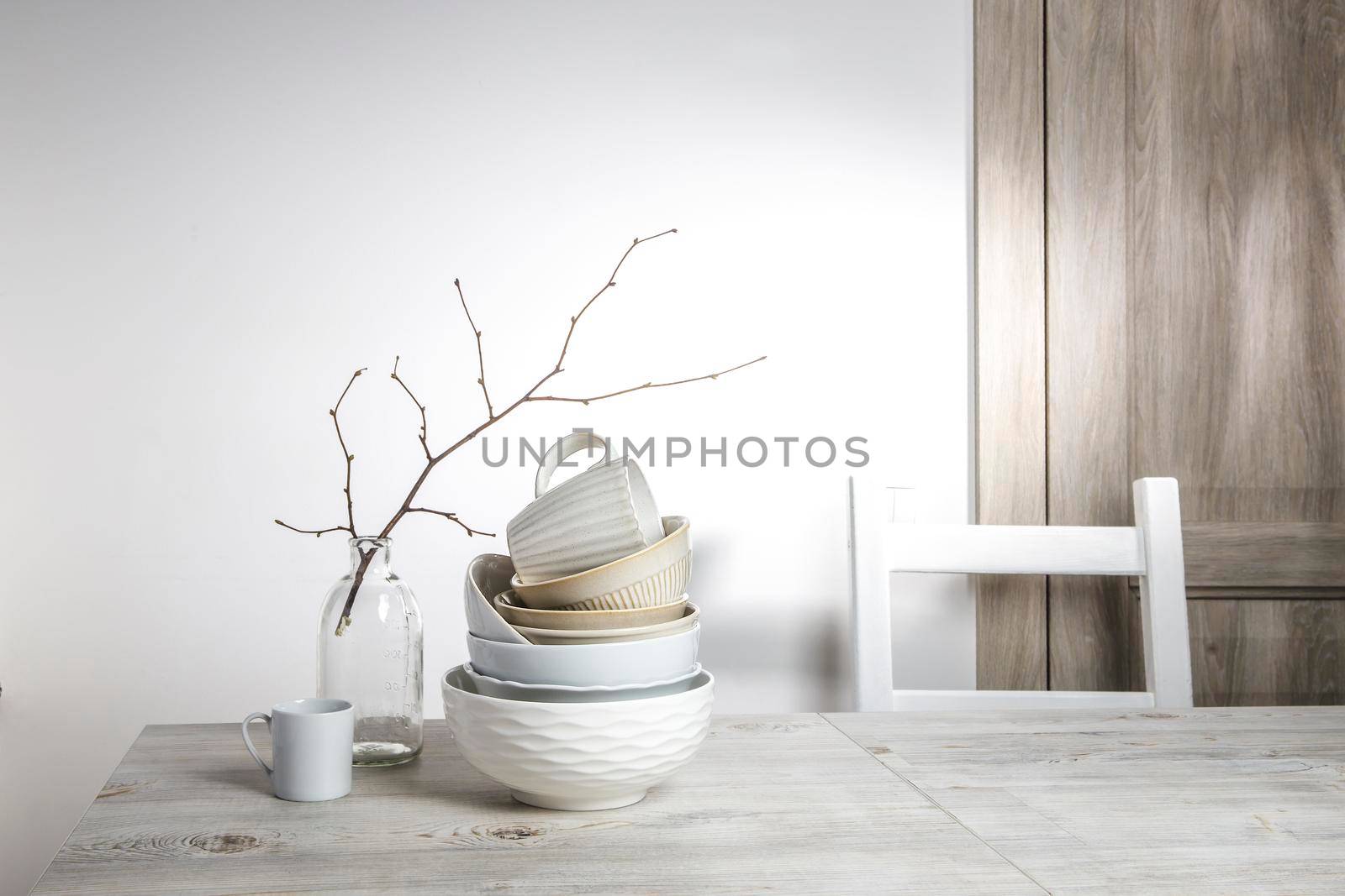 A pyramid of white bowls, plates and cups of different sizes and colors on a beige table in the kitchen. An unblown linden branch in a glass vase. Scandinavian style. Place for your text