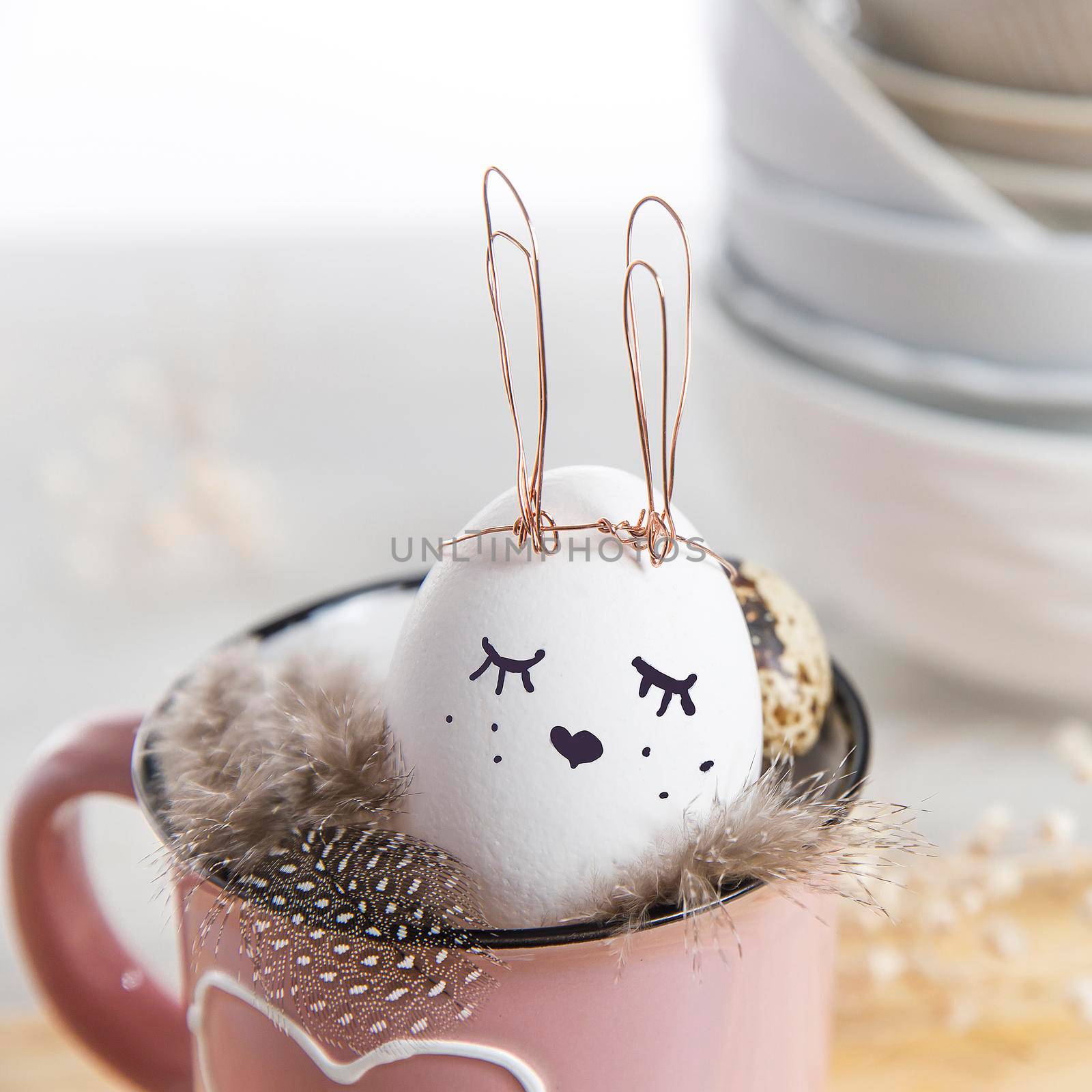 Easter concept. A white egg with copper wire ears and a painted rabbit sleeping face in a cup. Partridge eggs and feathers.