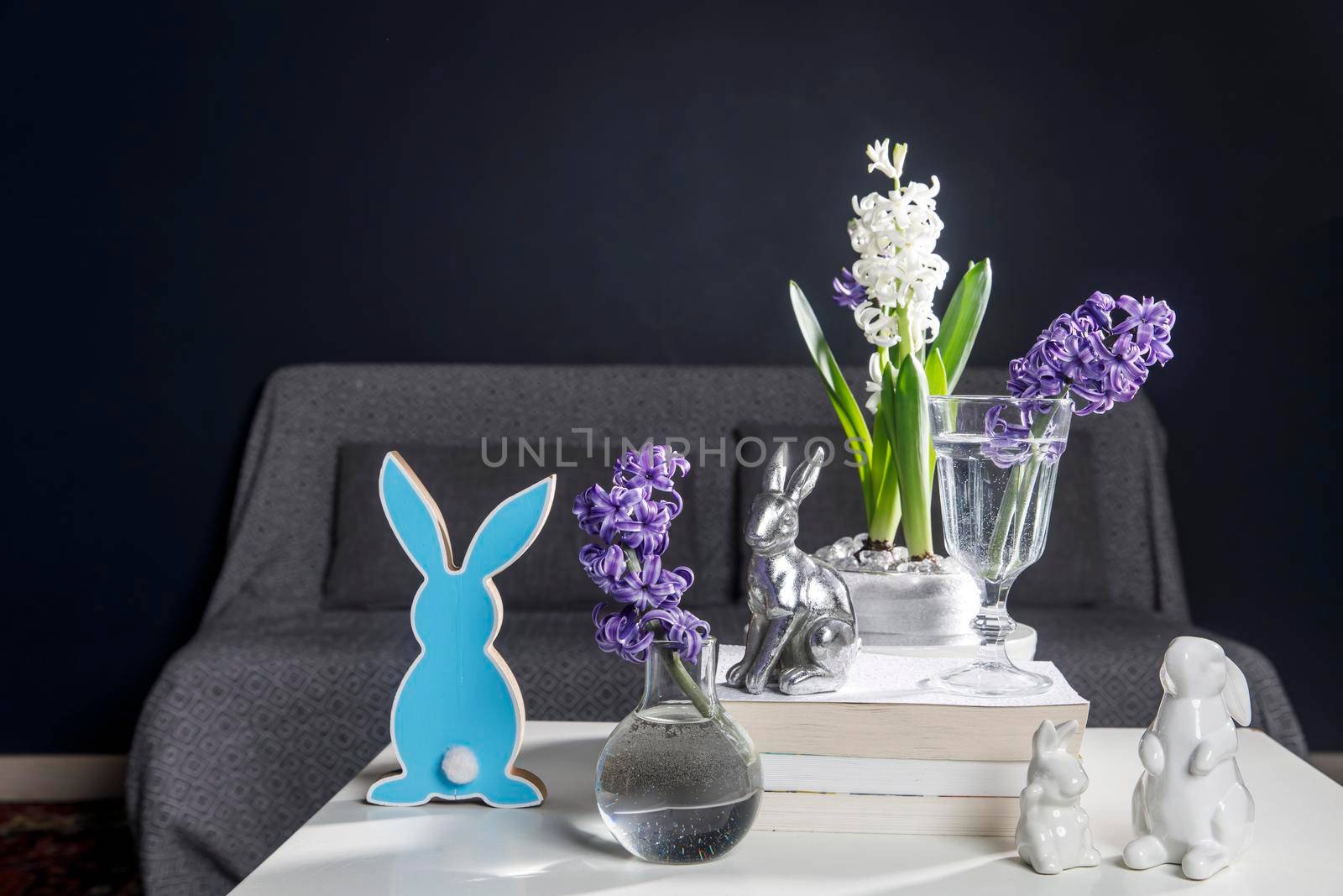Ceramic figurines of Easter bunnies of different sizes on the table. Blue and white hyacinths in glass cups on a dark background. Easter design by elenarostunova