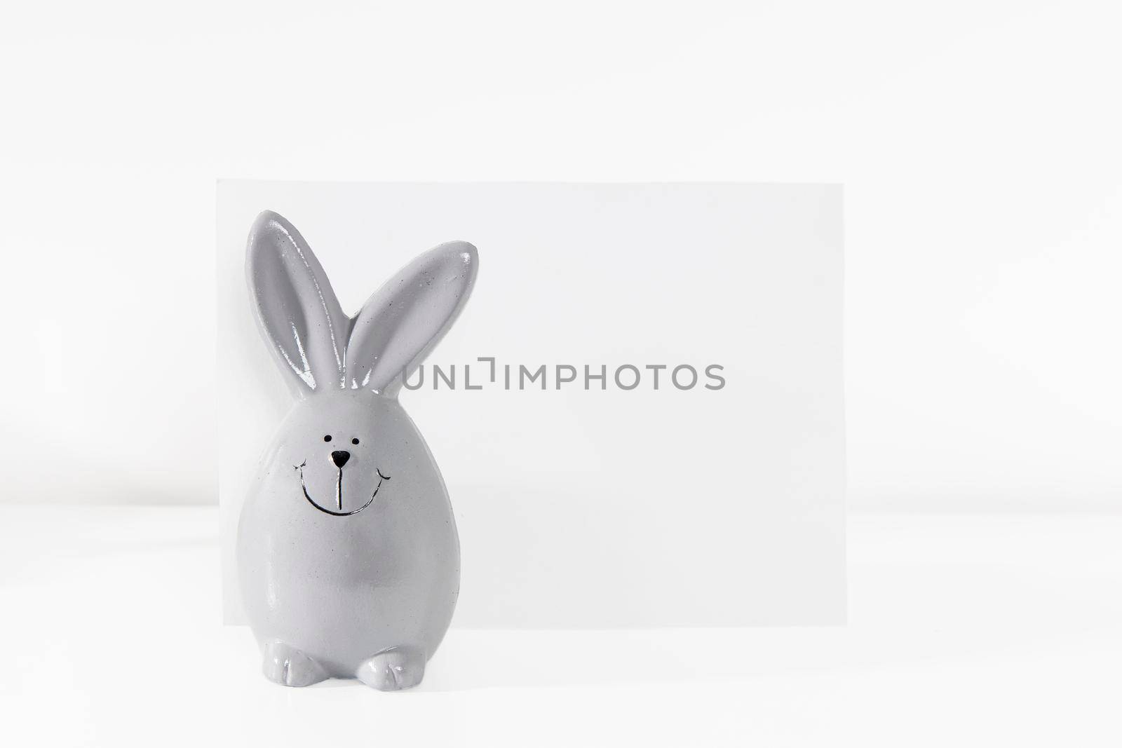 Mock up white frame with modern ceramic easter bunny decor with card on a shelf. White color scheme. Landscape frame orientation. by elenarostunova