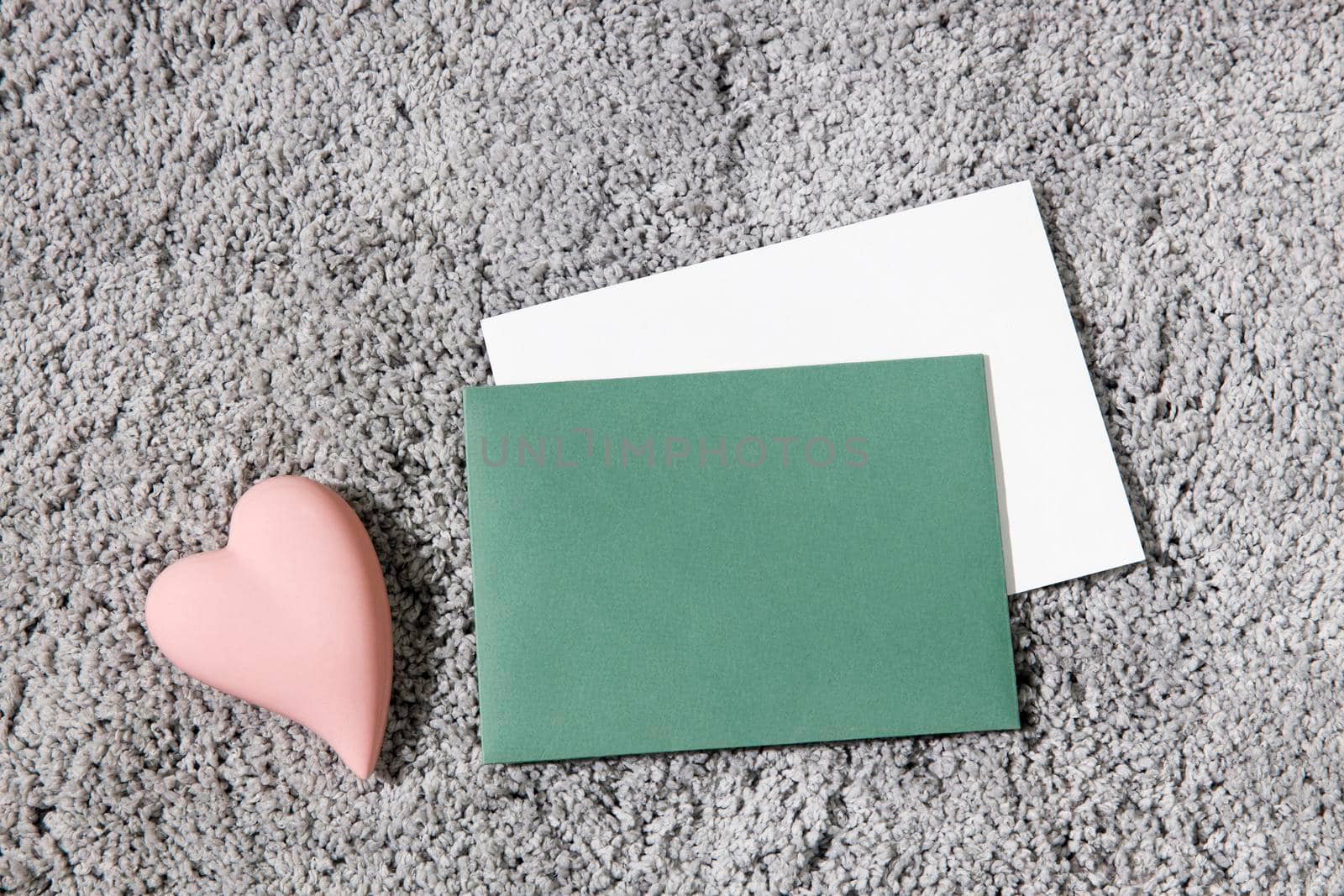 Greeting card for Valentine's Day. White and green envelopes, ceramic pink heart on a grey fluffy carpet background. Copy space. Place for text by elenarostunova