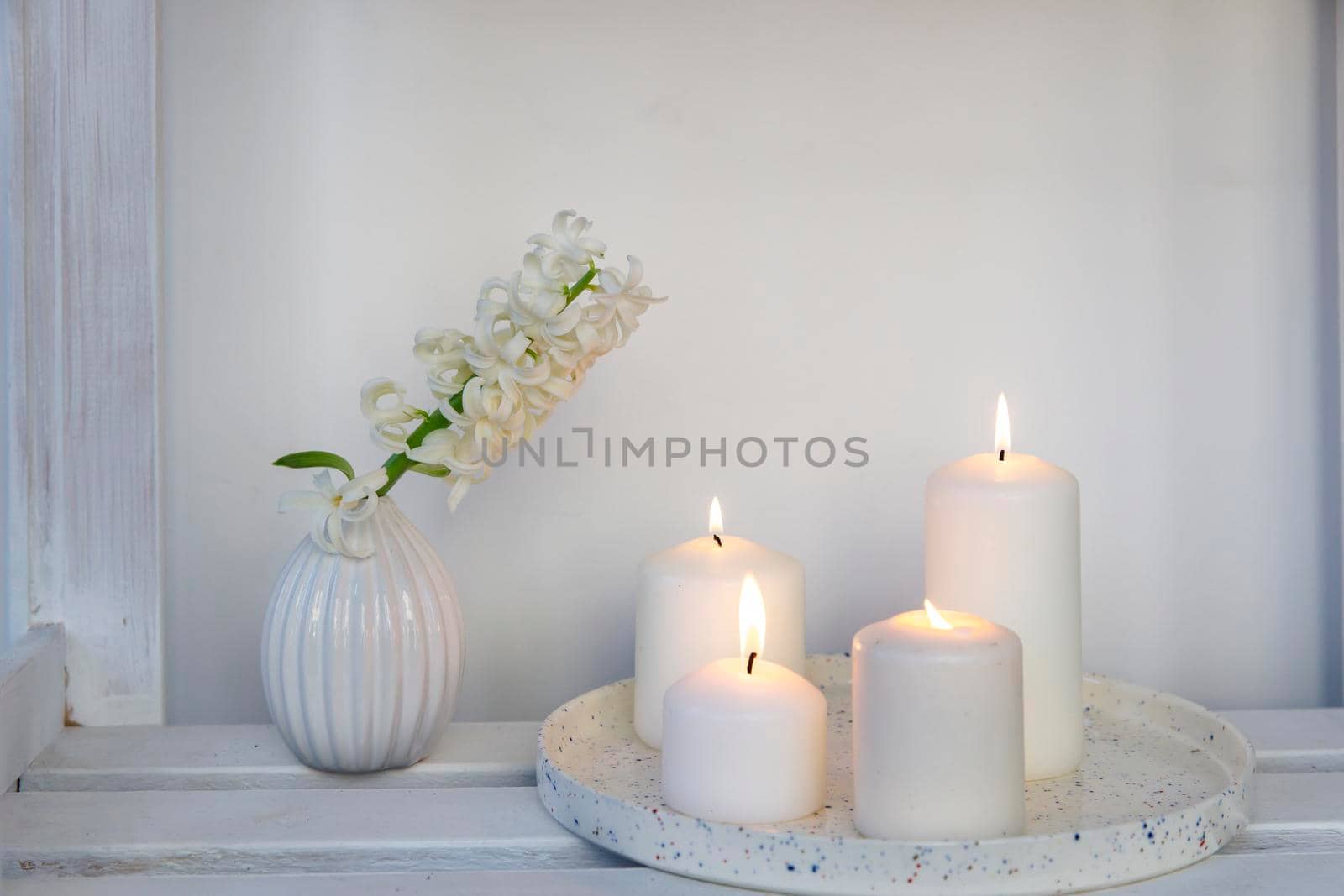 A white hyacinth in a 1970s fluted vase next to a tray of four lit candles is on the shelf. Minimalism. Scandinavian style.