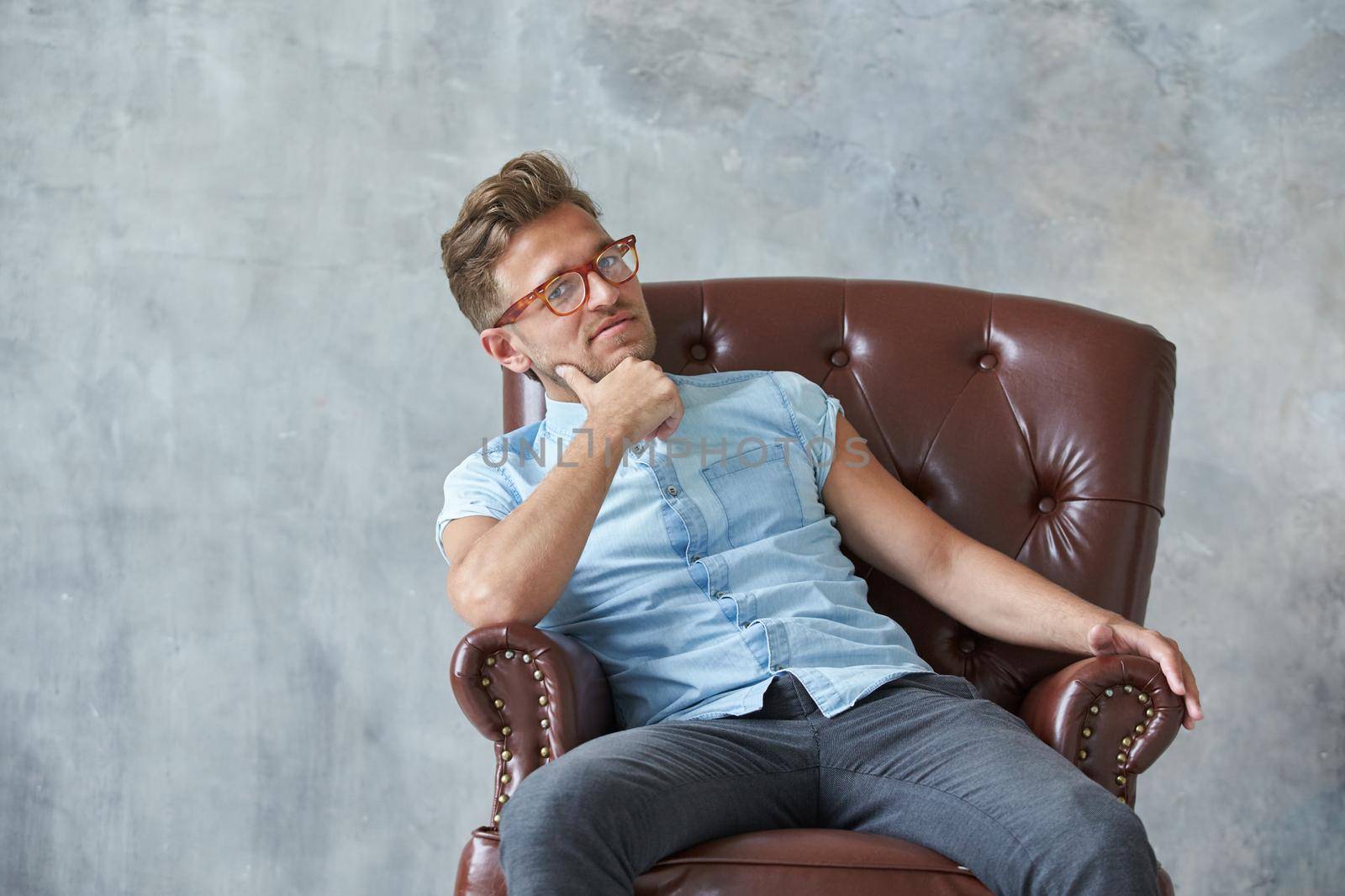 Portrait of a stylish intelligent man stares into the camera, small unshaven, charismatic, blue shirt, sitting on a brown leather chair, dialog, negotiation, short sleeve, brutal, hairstyle. High quality photo