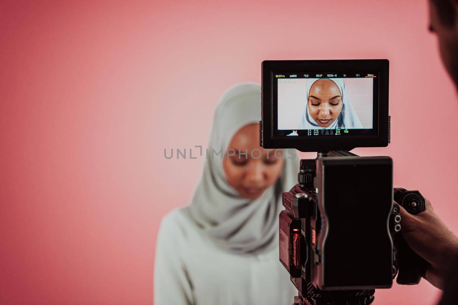 Videographer in digital studio recording video on professional camera by shooting female Muslim woman wearing hijab scarf plastic pink background. by dotshock