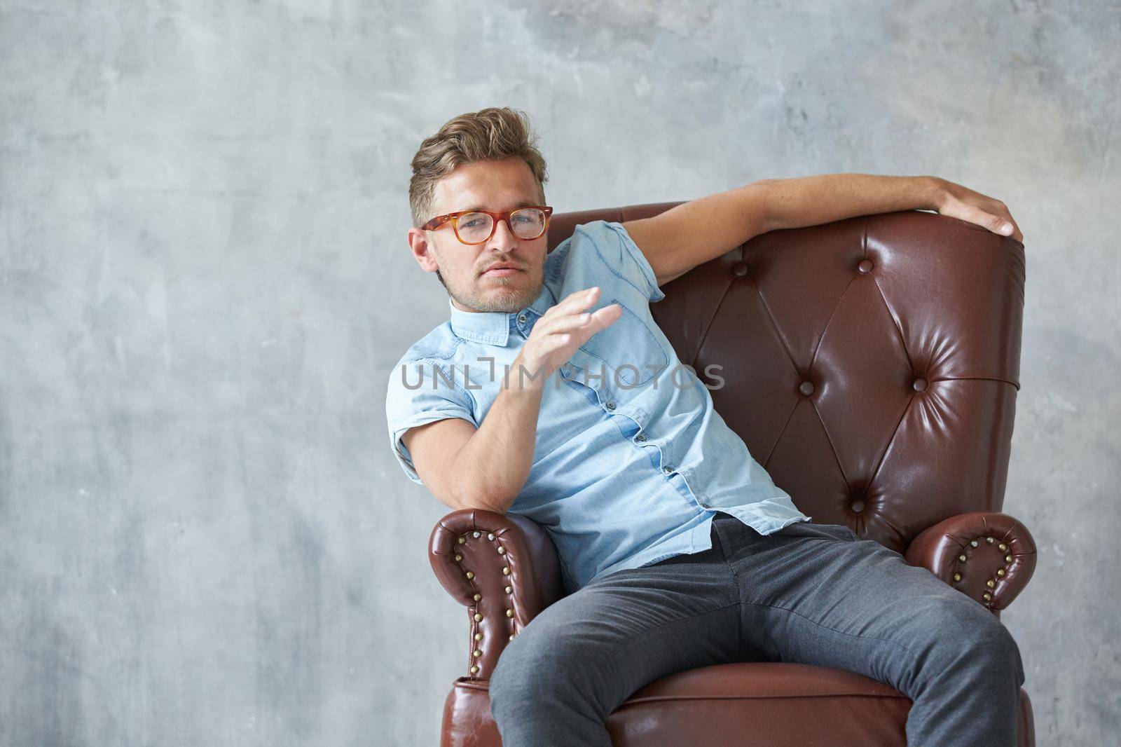 Portrait of a stylish intelligent man stares into the camera, small unshaven, charismatic, blue shirt, sitting on a brown leather chair, dialog, negotiation, short sleeve, brutal, hairstyle. High quality photo