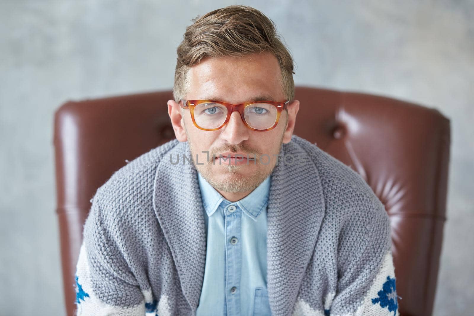 Portrait of a stylish intelligent man with glasses stares into the camera, good view, small unshaven, charismatic, blue shirt, gray sweater, sitting on a brown leather chair. High quality photo