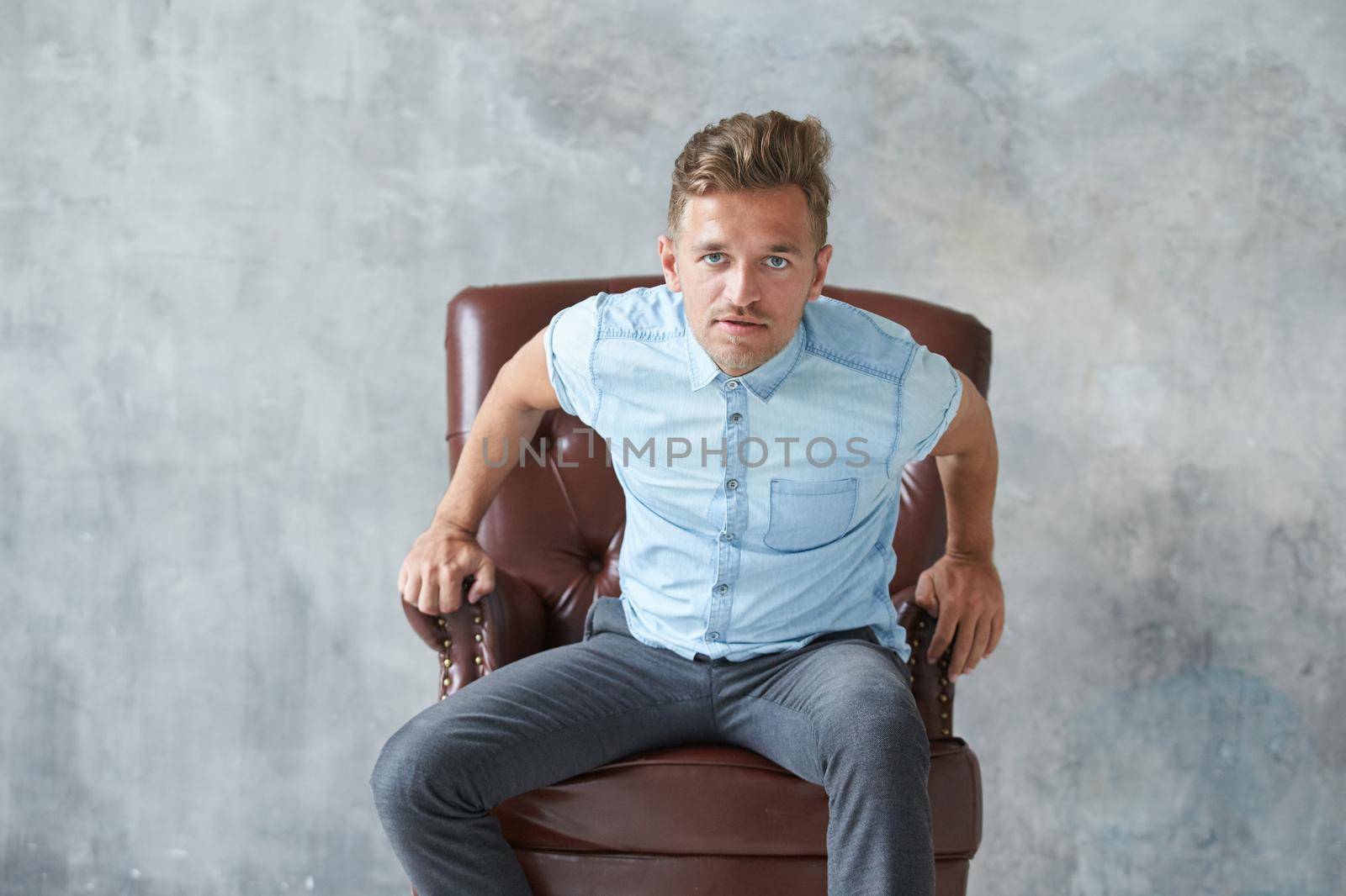 Portrait of a stylish intelligent man stares into the camera, small unshaven, charismatic, blue shirt, sitting on a brown leather chair, dialog, negotiation, short sleeve, brutal, hairstyle by vladimirdrozdin