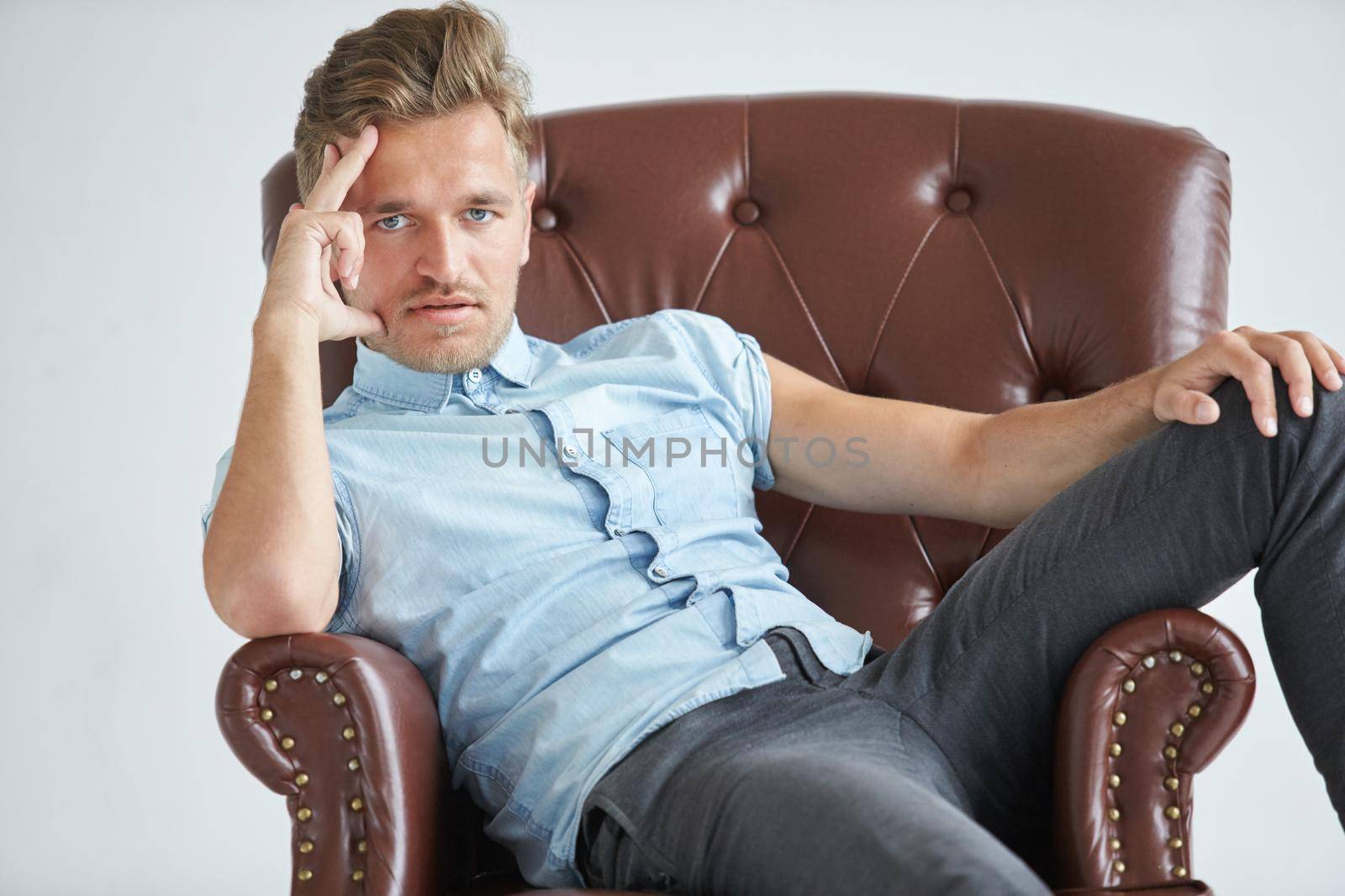 Portrait of a stylish intelligent man, small unshaven, charismatic, blue shirt, sitting on a brown leather chair, dialog, negotiation, short sleeve, brutal, hairstyle, emotions, energy, interest. High quality photo