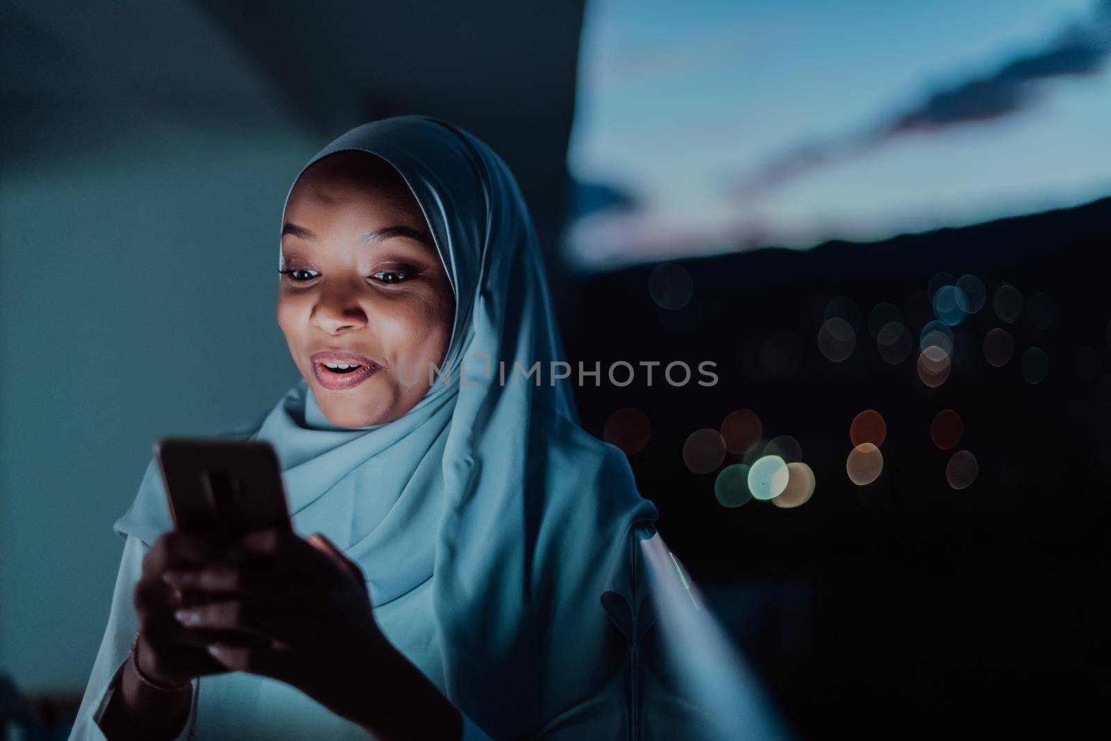 Young Muslim woman wearing scarf veil on urban city street at night texting on a smartphone with bokeh city light in the background. High-quality photo