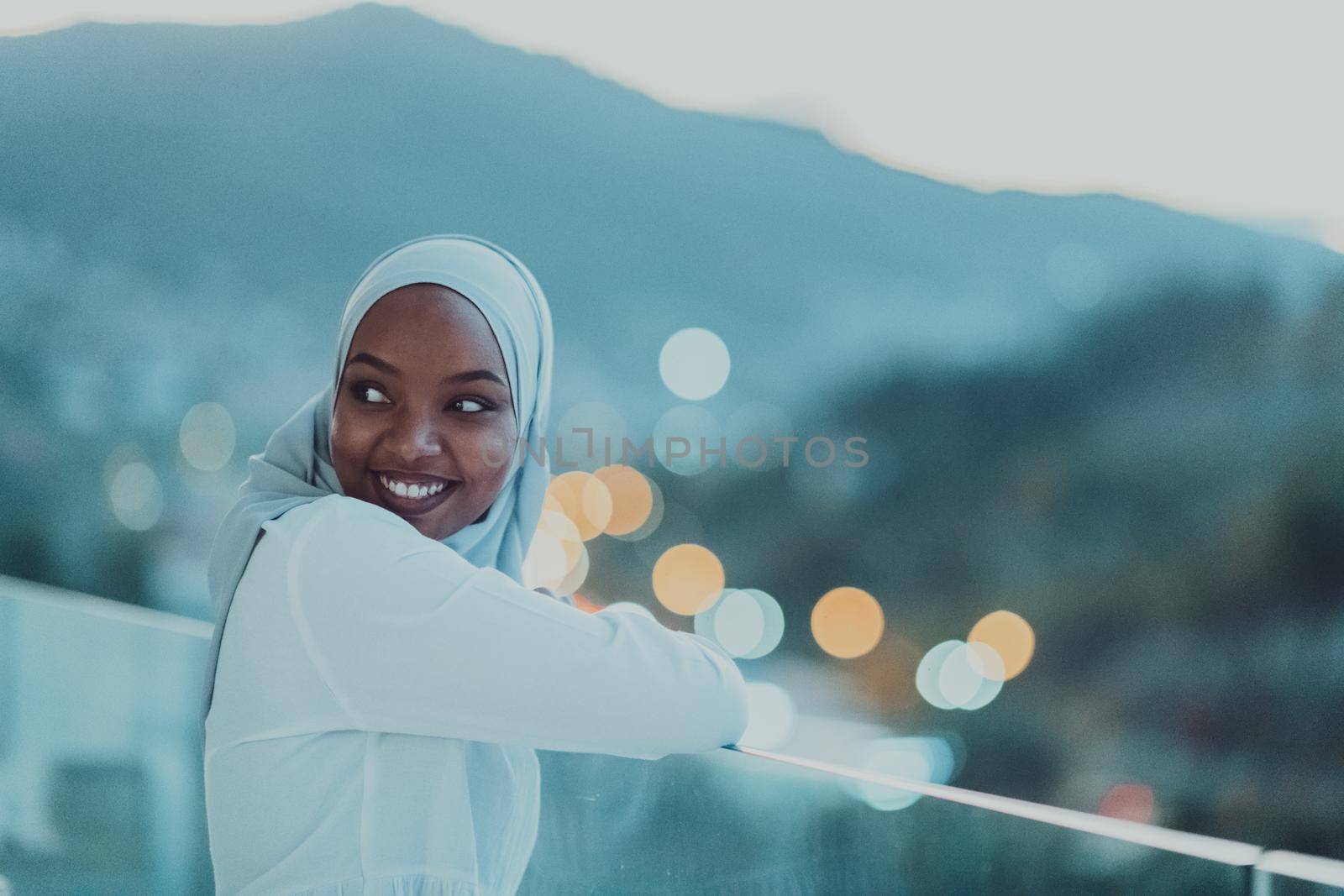 African Muslim woman in the night on a balcony smiling at the camera with city bokeh lights in the background. High-quality photo