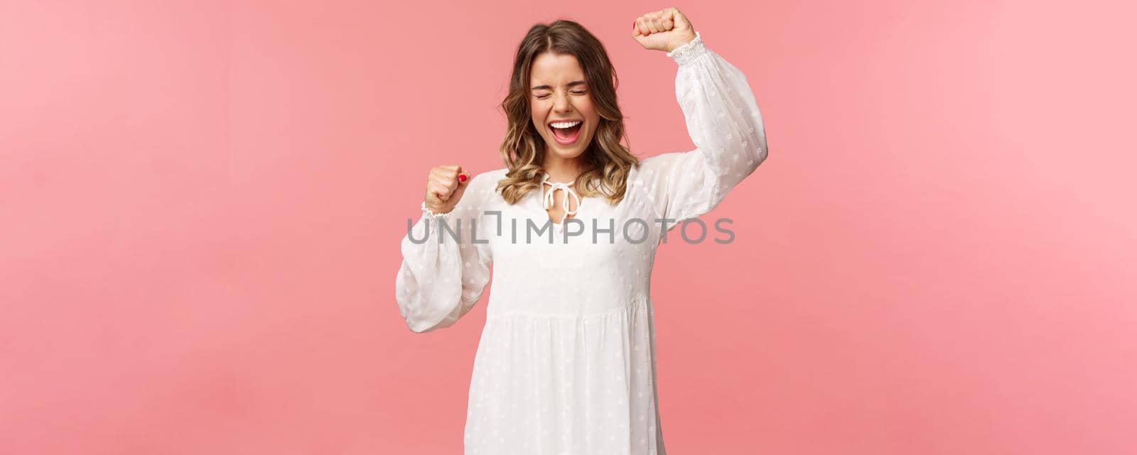 Portrait of beautiful tender young blond girl in white dress, raising hands up in hooray, yes or victory, shouting relieved, scream from happiness, triumphing, winning prize, become champion.
