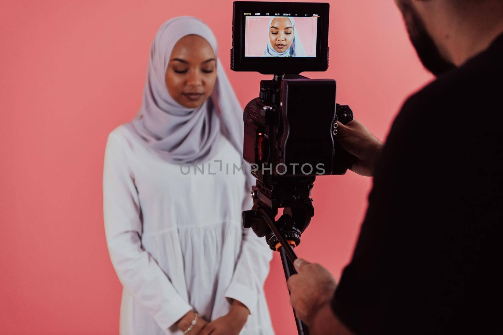 Videographer in digital studio recording video on professional camera by shooting female Muslim woman wearing hijab scarf plastic pink background. High-quality photo