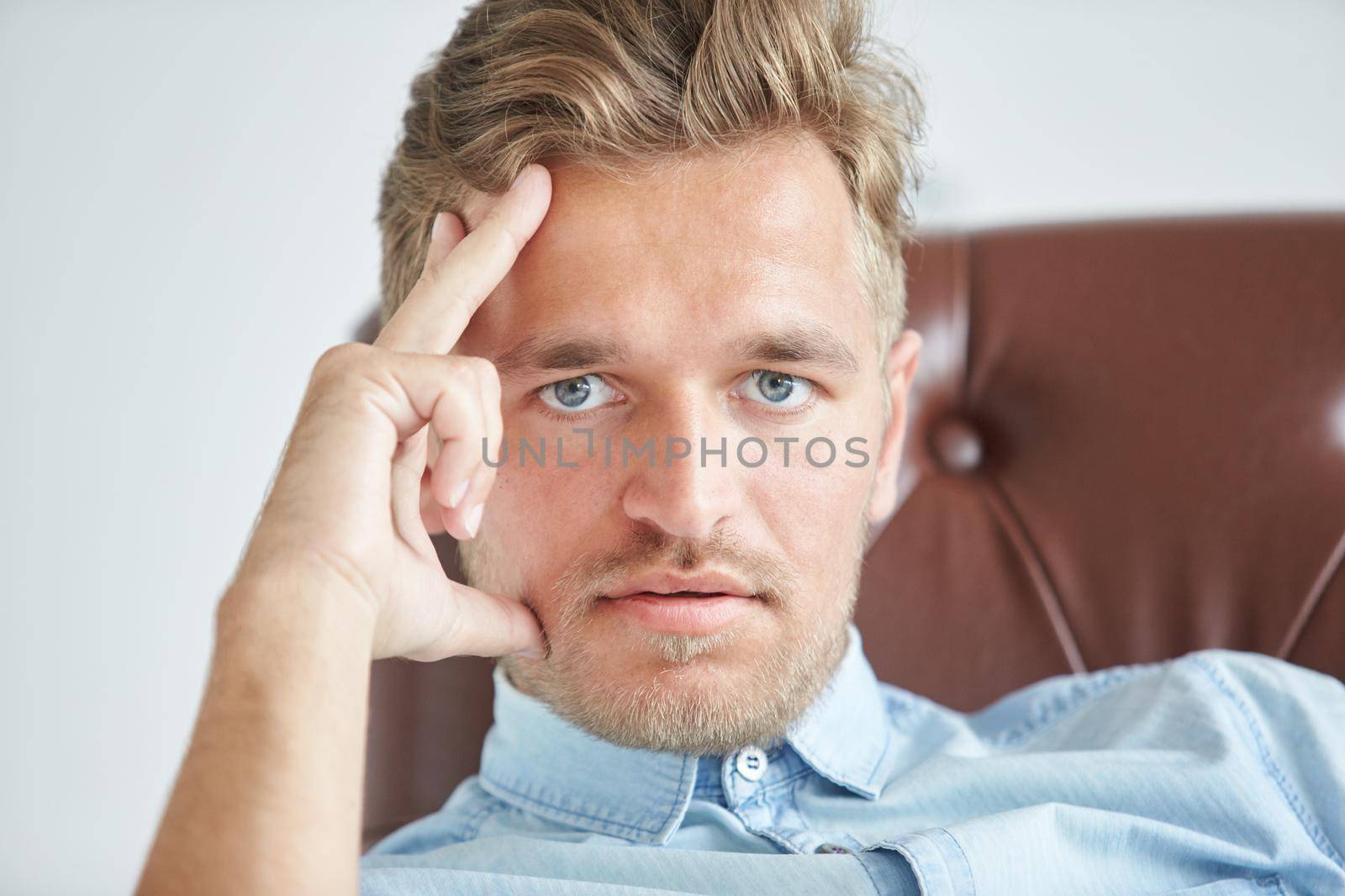 Portrait of a stylish intelligent man, small unshaven, charismatic, blue shirt, sitting on a brown leather chair, dialog, negotiation, short sleeve, brutal, hairstyle, emotions, energy, interest. High quality photo