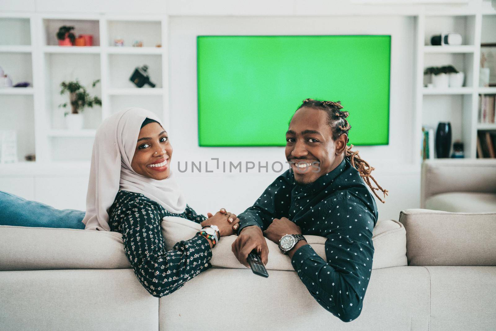 African Couple Sitting On Sofa Watching TV Together Chroma Green Screen Woman Wearing Islamic Hijab Clothes. High quality photo