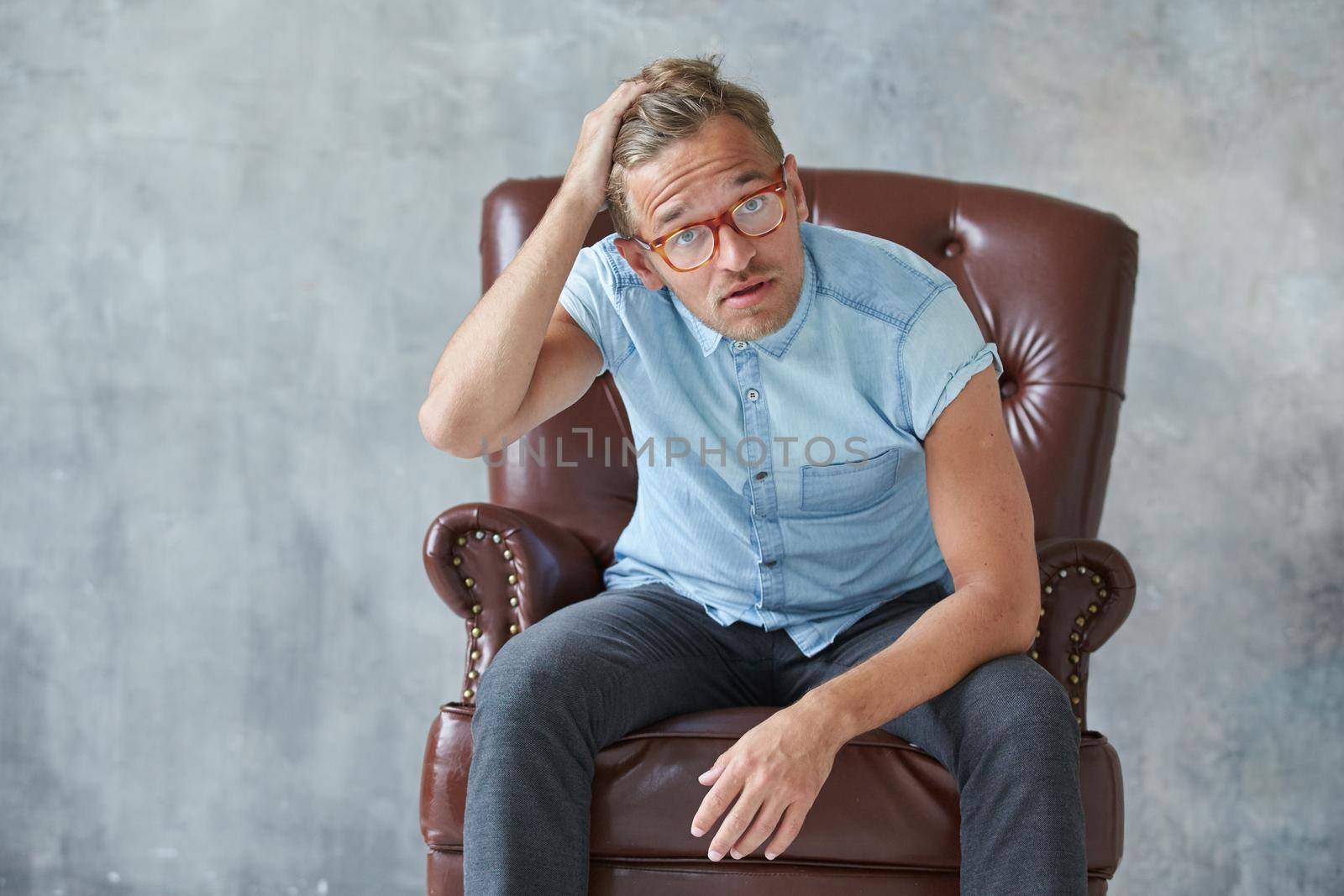 Portrait of a stylish intelligent man stares into the camera, small unshaven, charismatic, blue shirt, sitting on a brown leather chair, dialog, negotiation, short sleeve, brutal, hairstyle. High quality photo