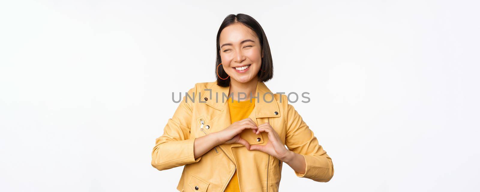 Image of beautiful asian girl showing heart, love sign, express care and romantic feelings, standing over white studio background by Benzoix