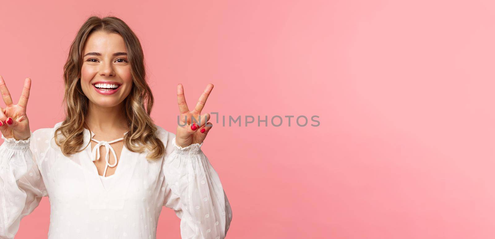 Close-up portrait of optimistic kawaii young blond girl with lovely beaming smile, showing peace signs and looking camera with positive attitude, enjoying spring, white dress, pink background.