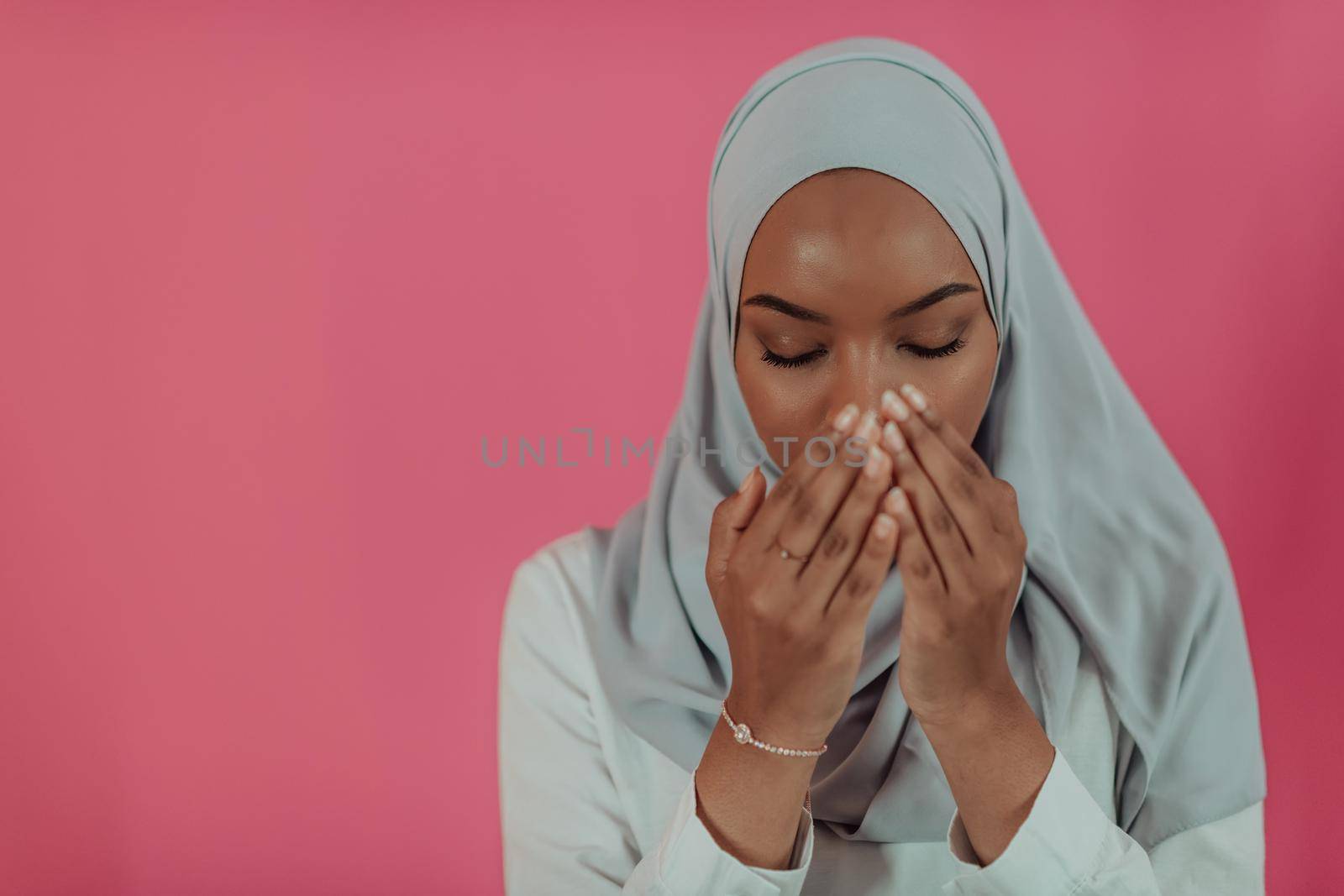 Modern African Muslim woman makes traditional prayer to God, keeps hands in praying gesture, wears traditional white clothes, has serious facial expression, isolated over plastic pink background. High-quality photo
