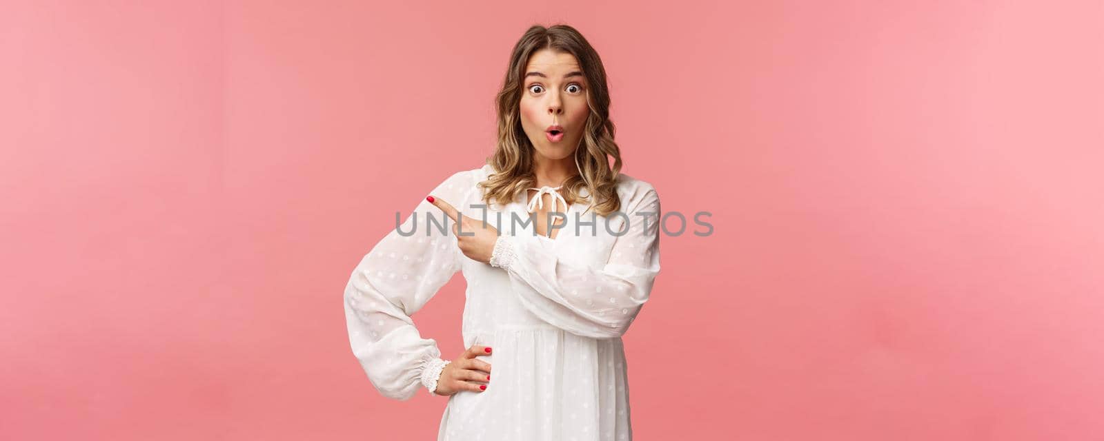 Fascinated and excited, curious blond caucasian girl in white dress, look intrigued at camera with folded lips, say wow and pointing finger left at blank space, standing pink background.