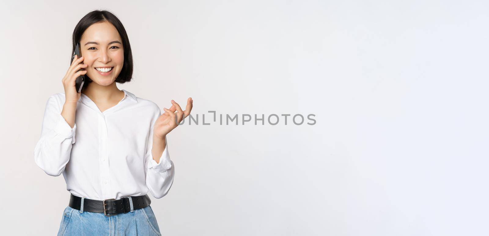 Smiling happy asian woman talking on smartphone with client, saleswoman on call, holding mobile phone and gesturing, standing over white background.