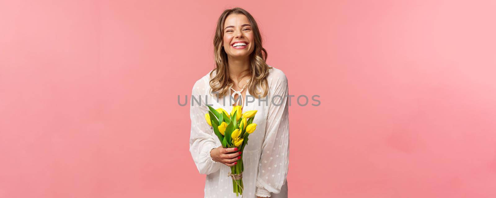 Holidays, beauty and spring concept. Carefree happy attractive blond girl in white dress holding yellow tulips and laughing with pleased expression, standing pink background, have romantic date by Benzoix