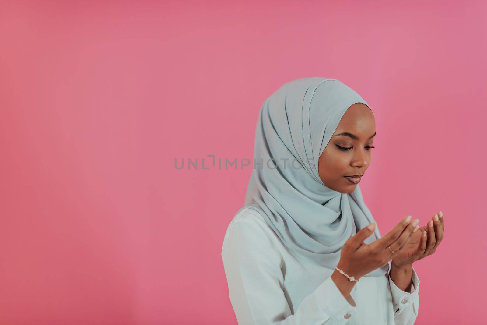 Modern African Muslim woman makes traditional prayer to God, keeps hands in praying gesture, wears traditional white clothes, has serious facial expression, isolated over plastic pink background. High-quality photo