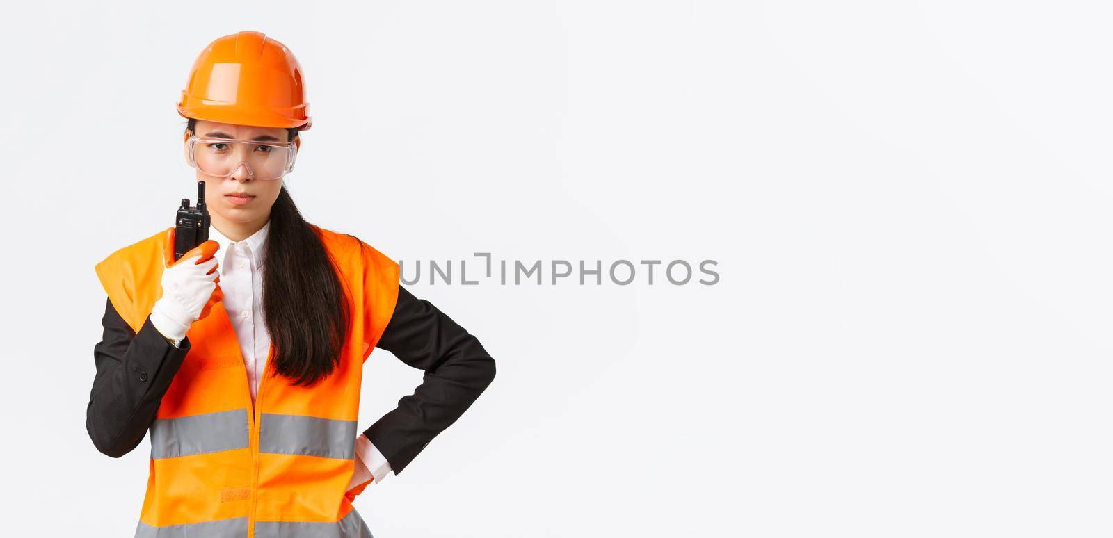 Disappointed asian female construction engineer, technician or industrial manager in safety uniform calling employee via walkie-talkie, scolding personal using radio communication at enterprise.