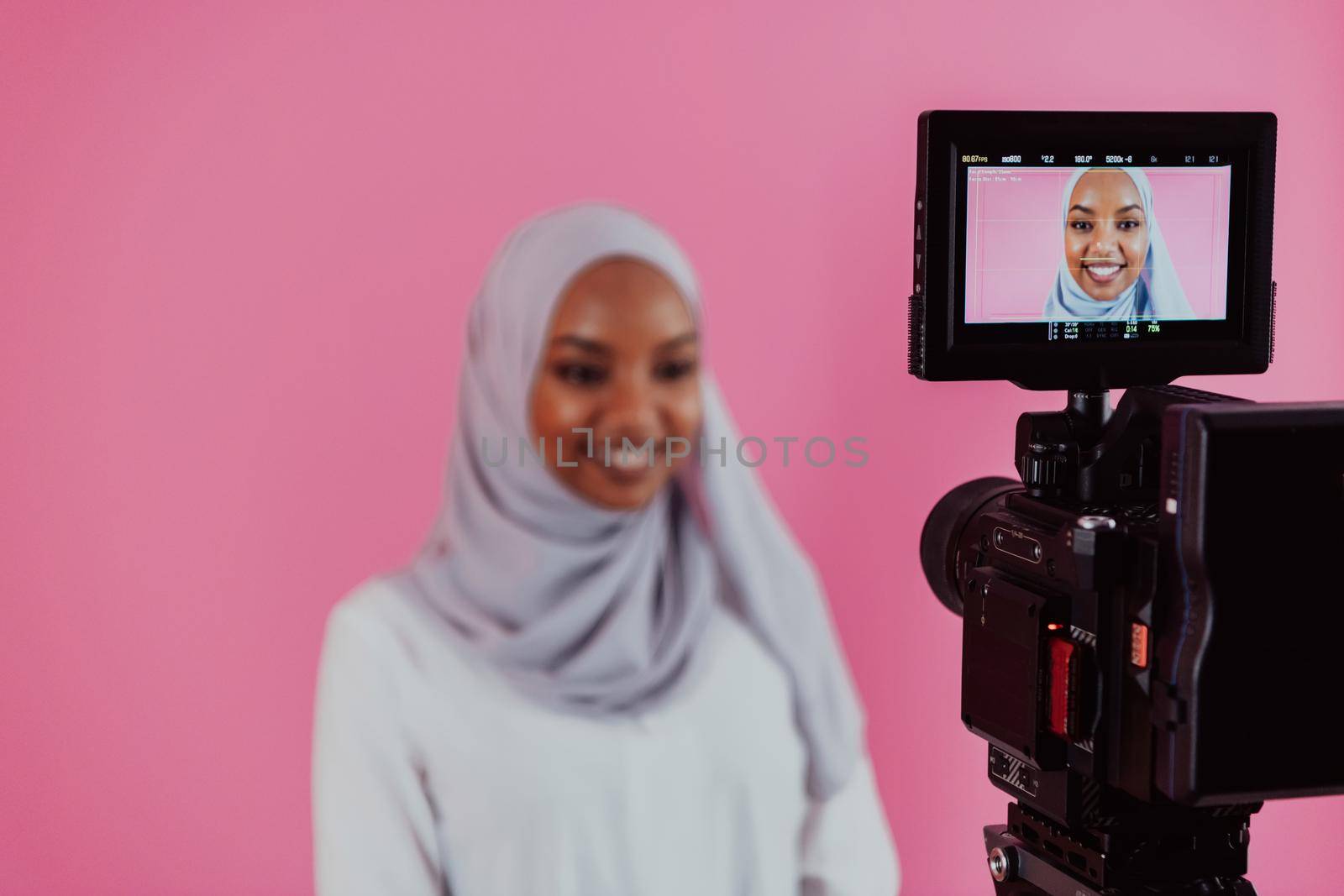 Videographer in digital studio recording video on professional camera by shooting female Muslim woman wearing hijab scarf plastic pink background. High-quality photo