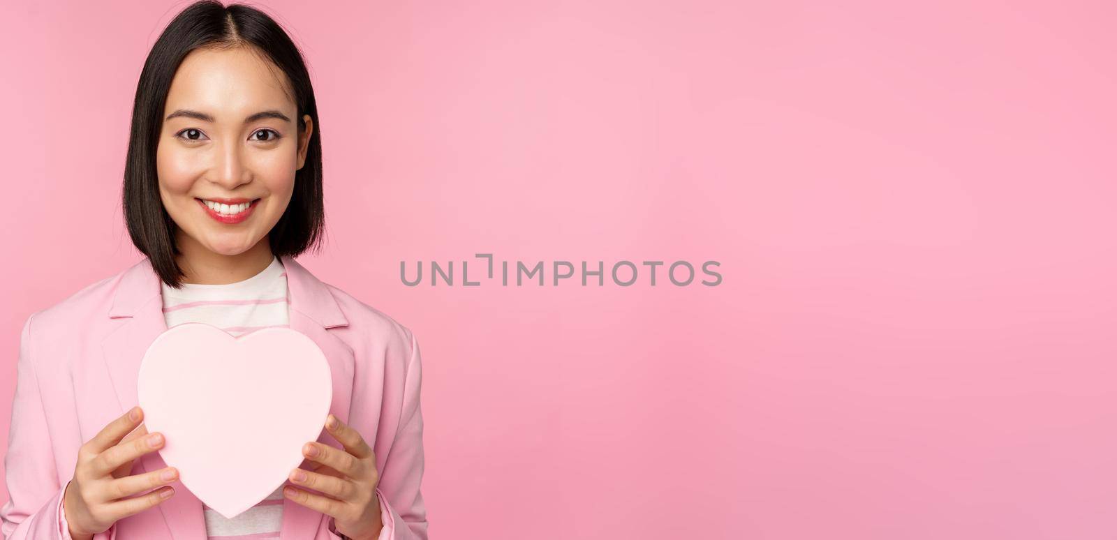 This is for you. Romantic cute asian corporate woman, girl in suit, showing heart-shaped box with gift, standing over pink background.