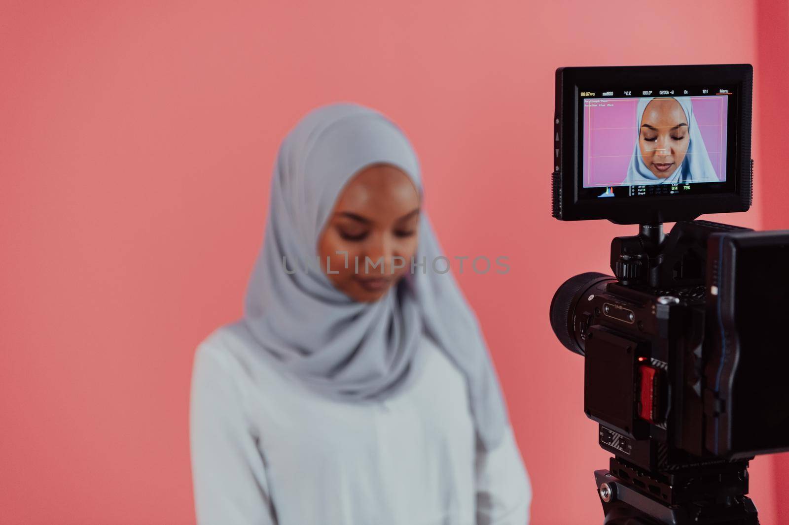 Videographer in digital studio recording video on professional camera by shooting female Muslim woman wearing hijab scarf plastic pink background. High-quality photo