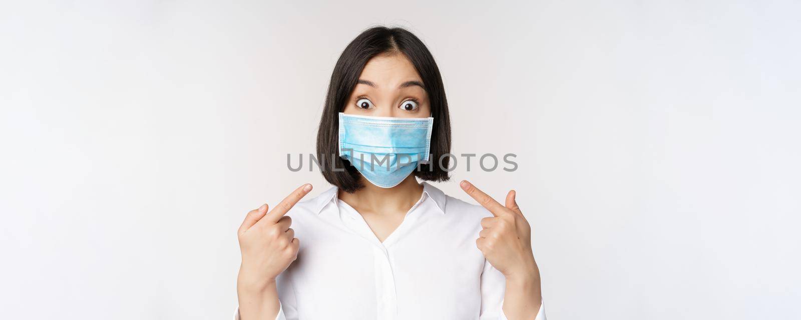 Image of amazed young asian woman pointing at herself while wearing medical face mask, standing over white background.