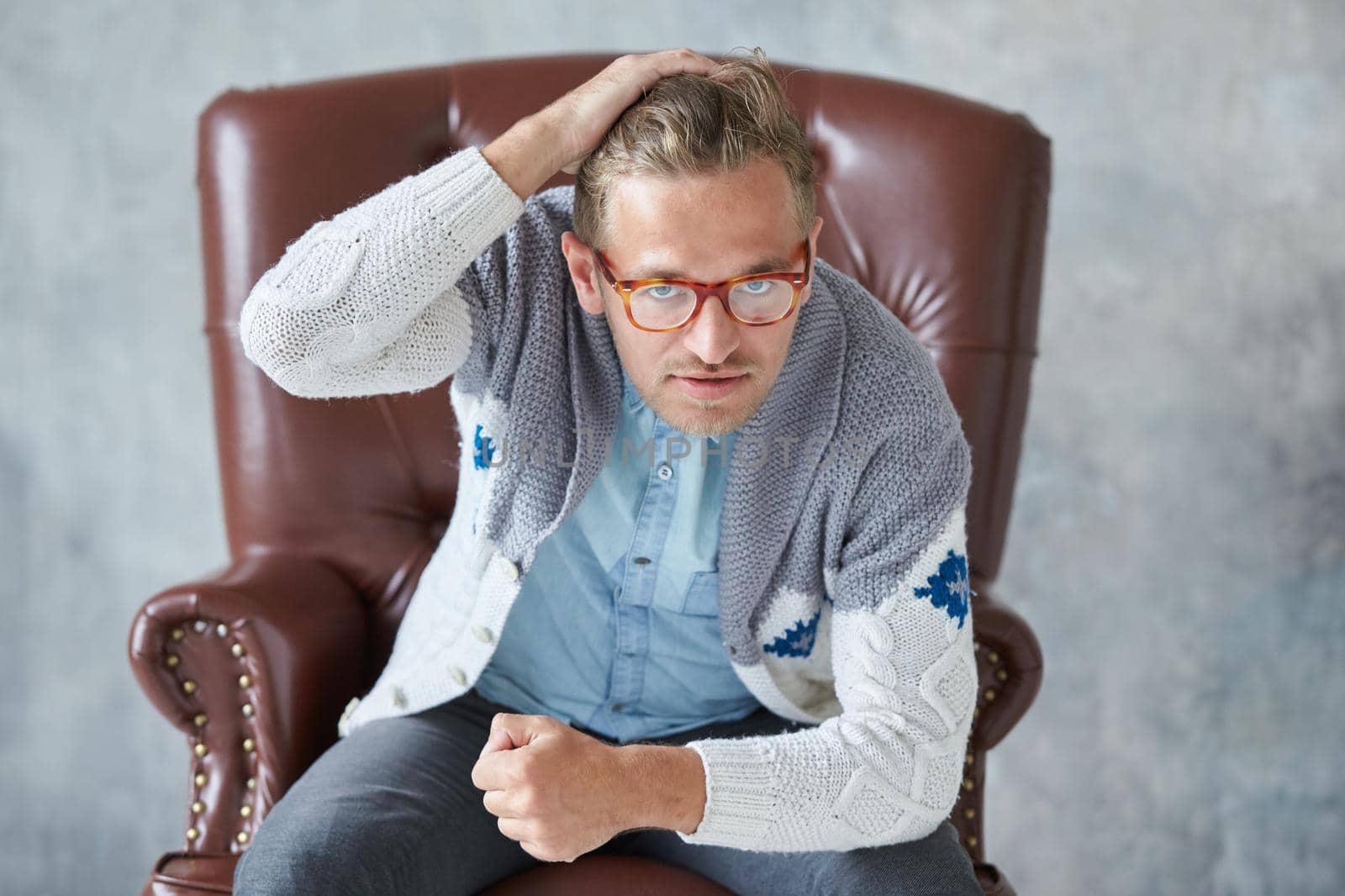 Portrait of a stylish intelligent man with glasses stares into the camera, good view, small unshaven, charismatic, blue shirt, gray sweater, sitting on a brown leather chair. High quality photo