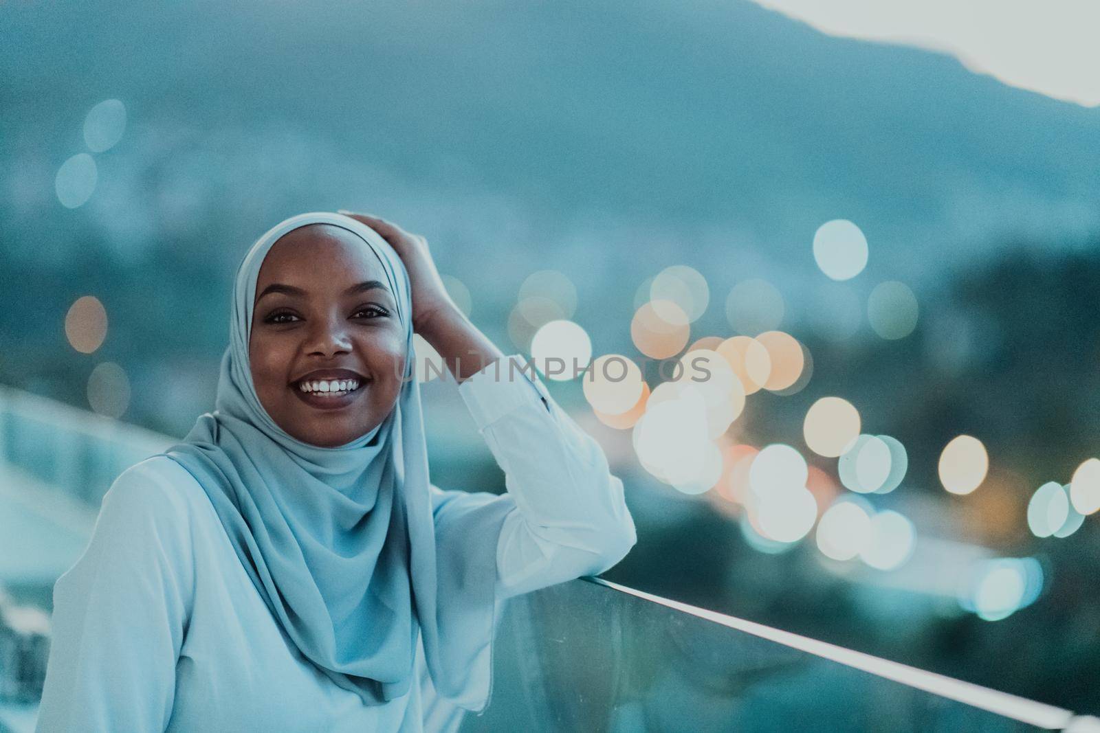 African Muslim woman in the night on a balcony smiling at the camera with city bokeh lights in the background. High-quality photo