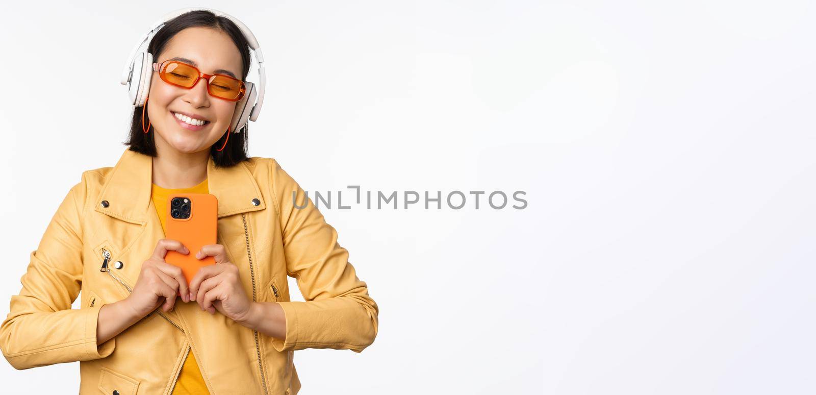 Technology concept. Stylish asian girl in headphones, holding smartphone, dancing and singing, listening music, standing over white background.