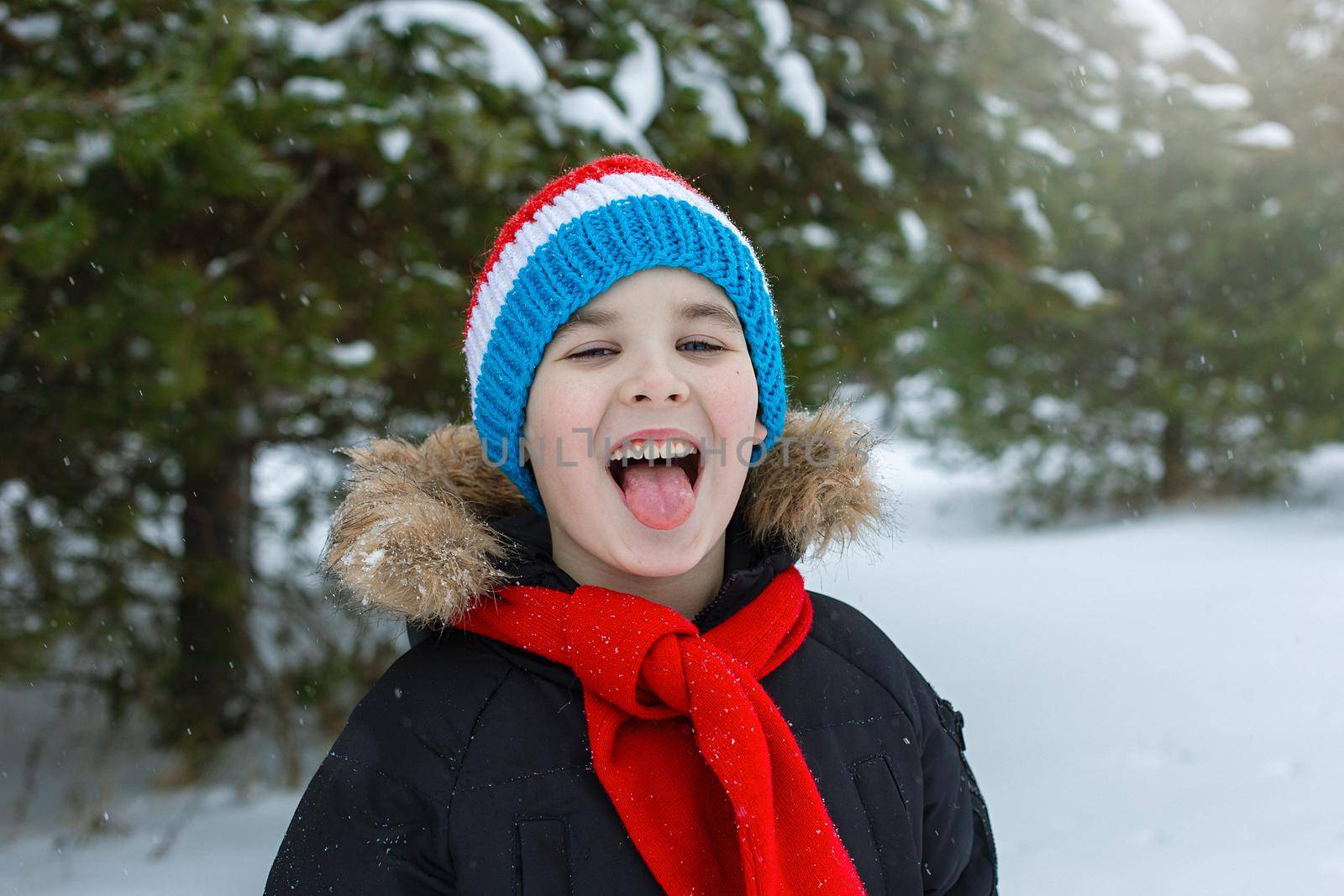Funny boy in bright winter clothes and a red scarf walks during a snowfall, catches snowflakes with his tongue
