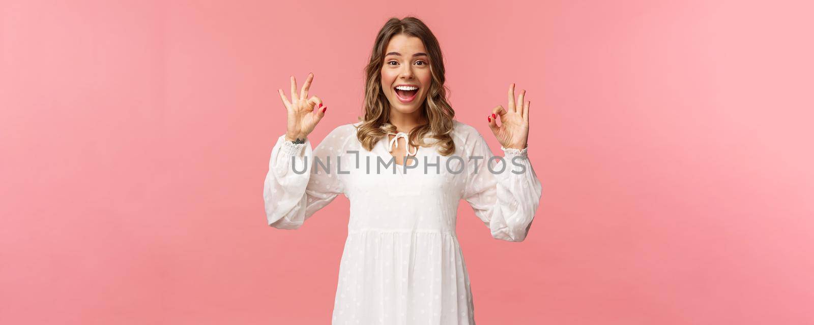 Portrait of happy and pleased good-looking blond woman in white dress, recommend great product, leave positive review, show okay signs and smiling satisfied, standing pink background.