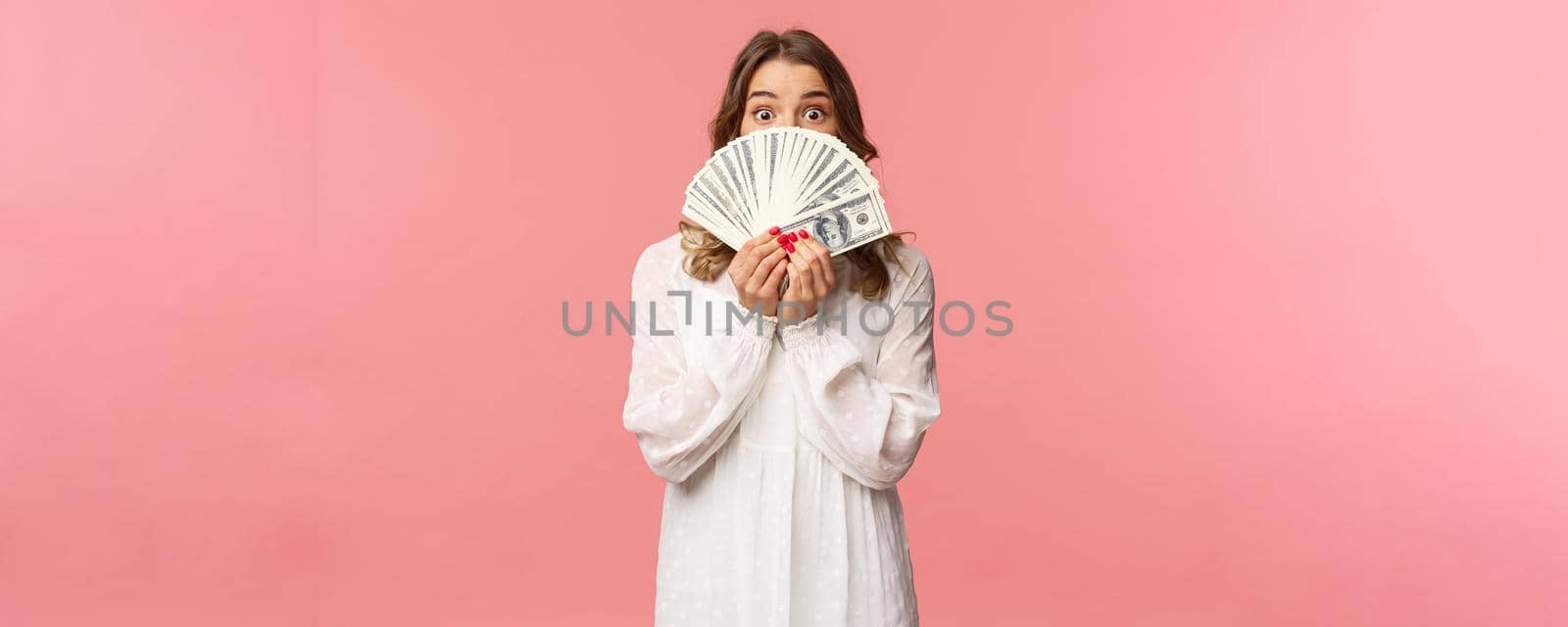 Portrait of amazed and excited cute feminine blond girl in white dress, holding dollars over face, looking from under cash at camera with surprised expression, stand pink background by Benzoix