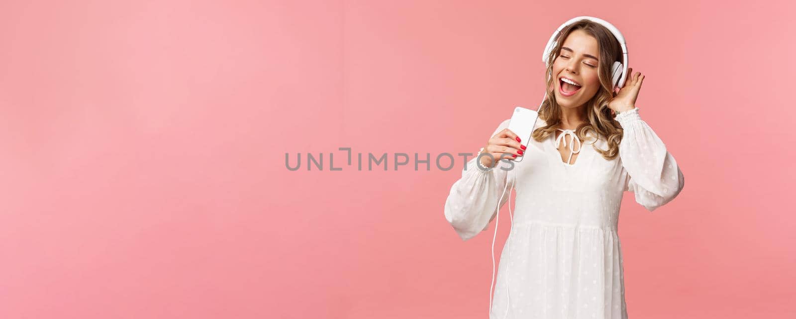 Portrait of beautiful blond caucasian female in white dress, listening music in headphones, close eyes and smiling, using mobile phone like microphone, singing along favorite song, pink background.