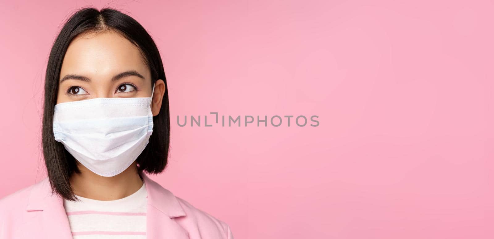 Close up portrait of japanese corporate woman in medical face mask from covid-19, looking left at logo, sale promo, standing over pink background.