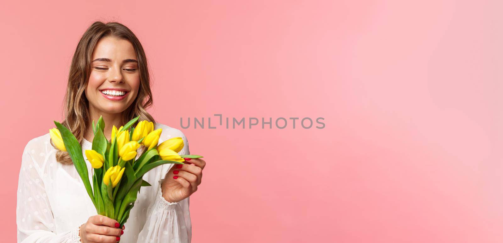 Spring, happiness and celebration concept. Close-up portrait of lovely romantic smiling girl touching petal of yellow tulip, holding flowers, receive bouquet on date, standing pink background.